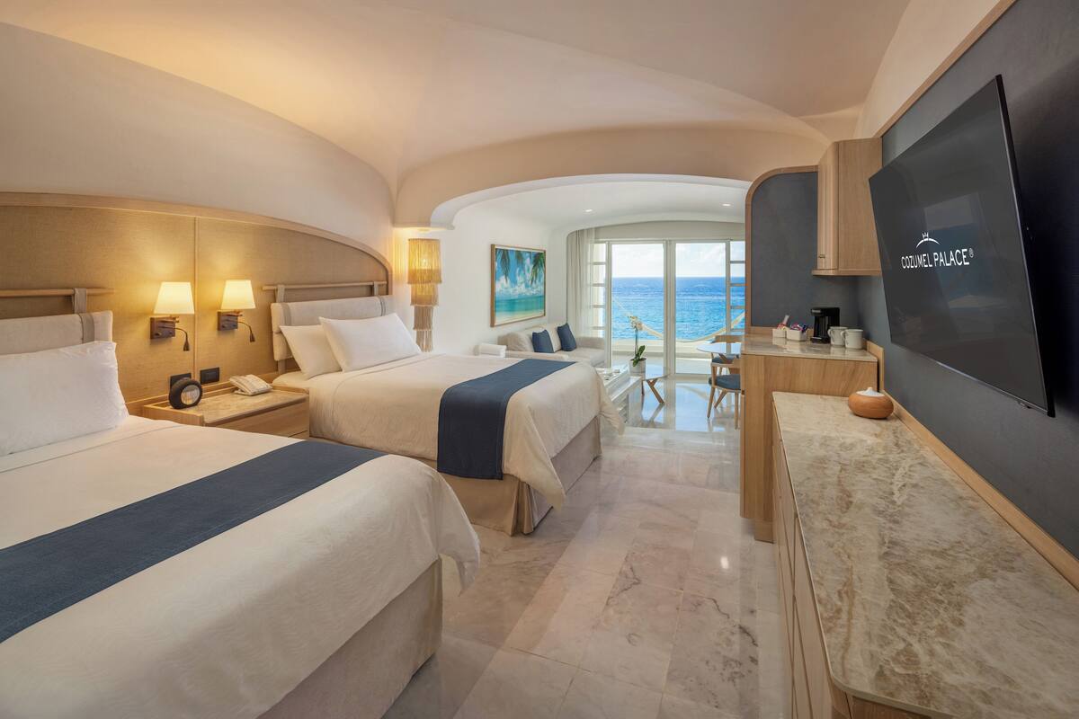A hotel room with two beds, a desk, a TV, and a view of the ocean.