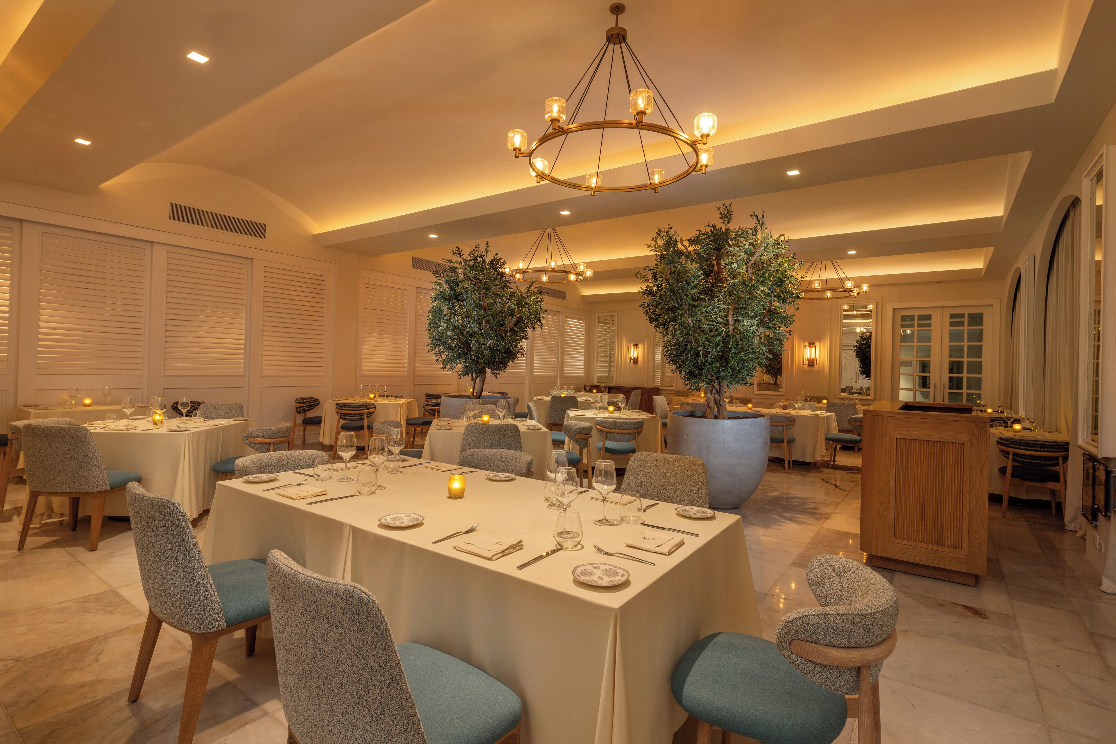 A spacious dining area with multiple tables and chairs, a chandelier, and potted plants.