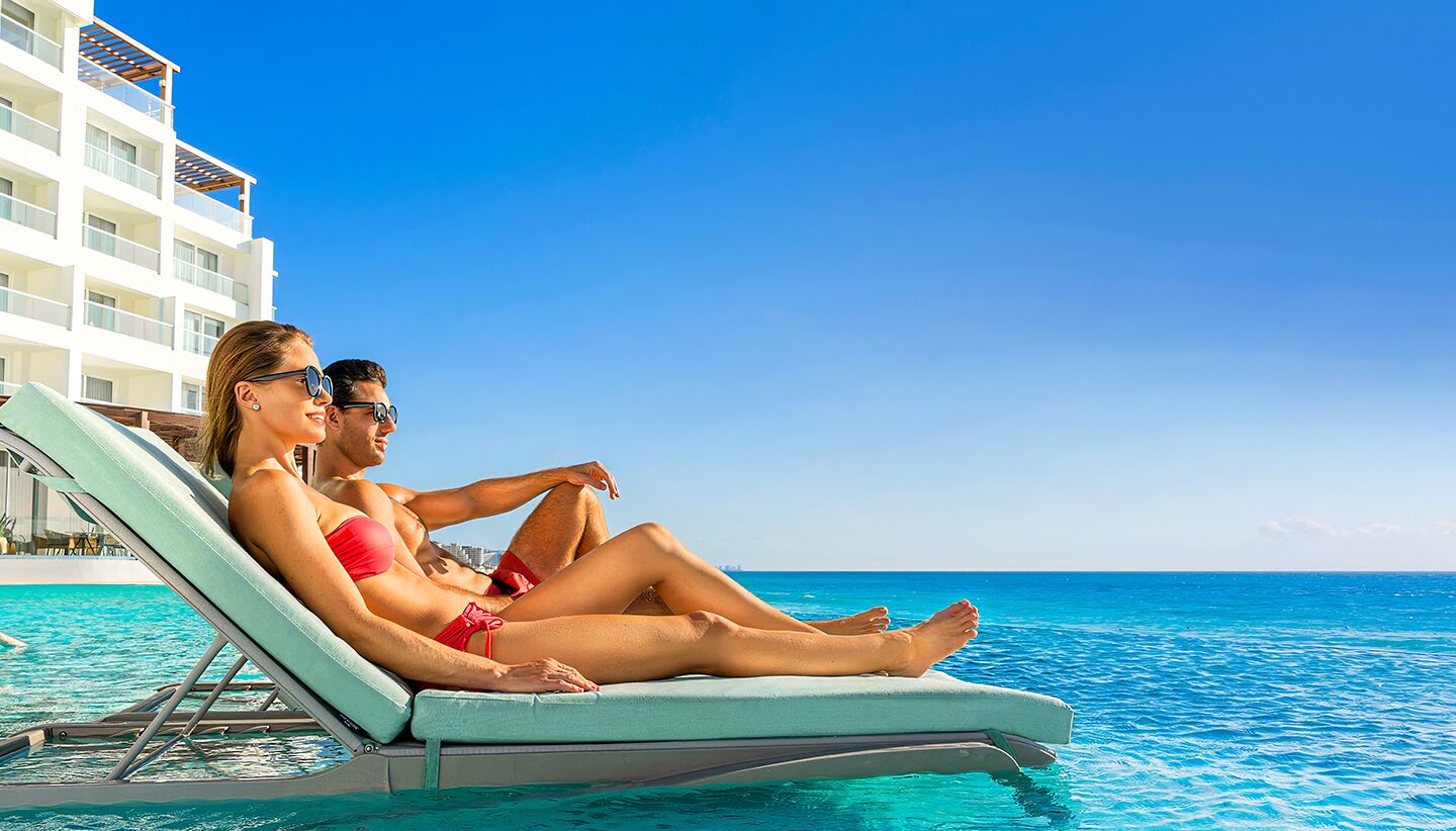 A couple relaxing on a poolside lounge chair with a view of the ocean and a hotel building in the background.