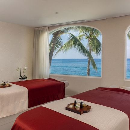 A room with two beds, each with a red bedspread and a tray of toiletries. There is a large window with a view of the ocean and palm trees.
