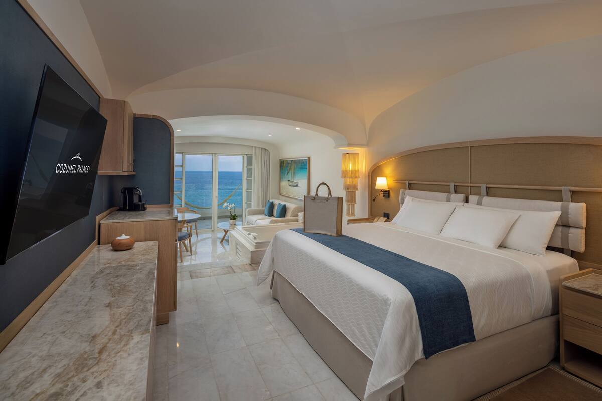 A modern hotel room with a large bed, a desk, a TV, and a view of the sea.