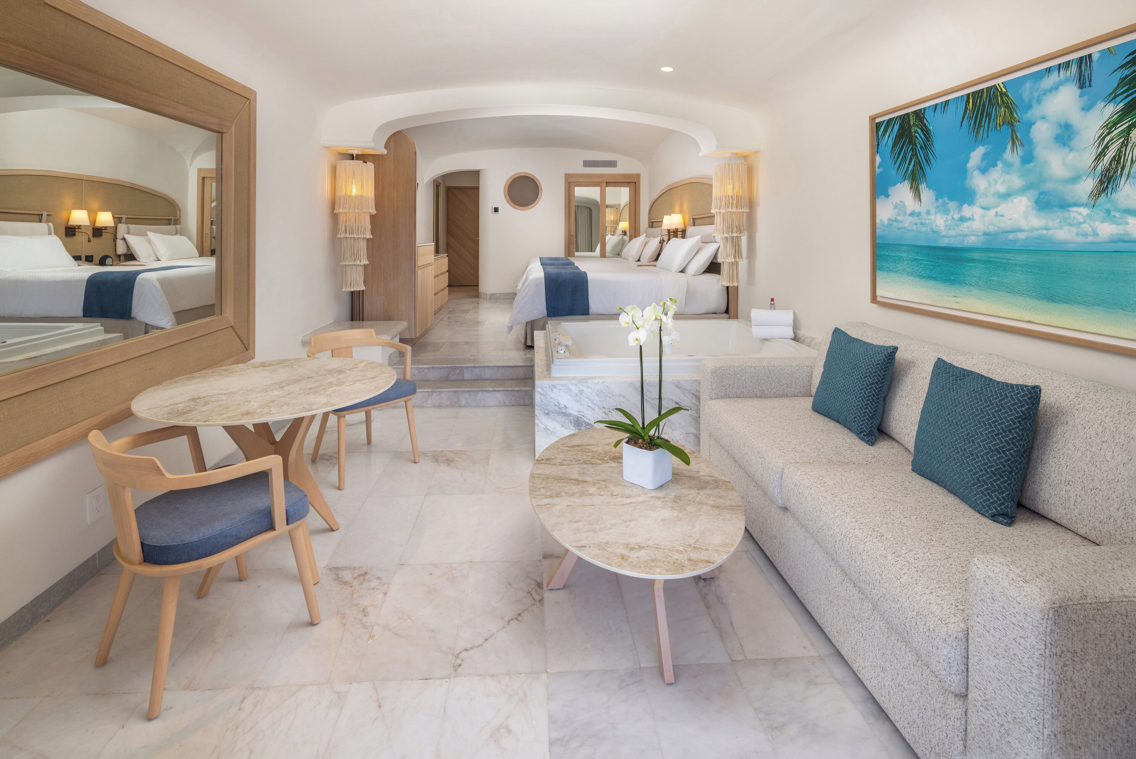 A hotel room with a bed, a sofa, a dining table with chairs, a coffee table, and a large painting of a beach.