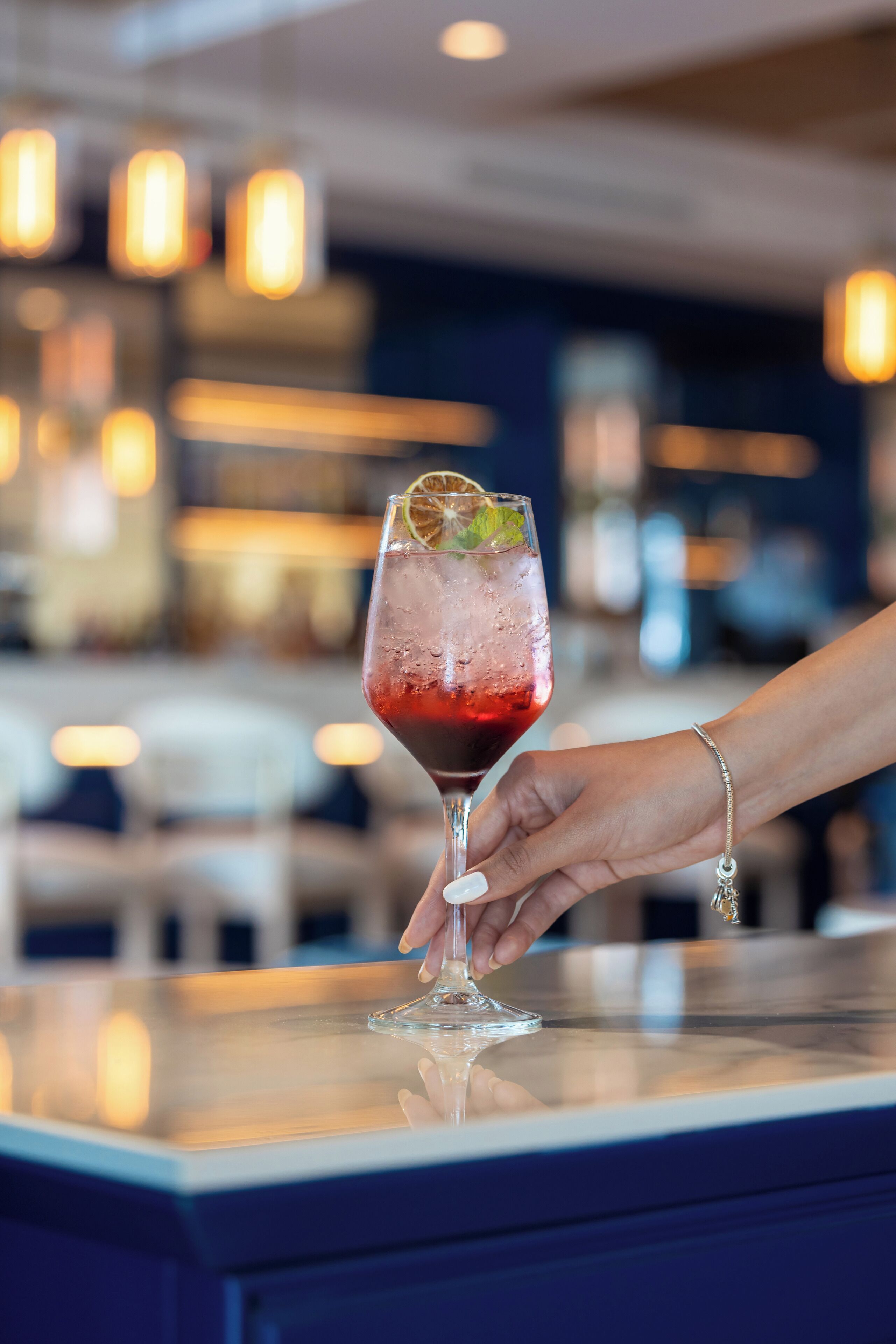 Eine Hand hält ein Glas mit einem roten Getränk, verziert mit einer Limettenscheibe, auf einer Bar.