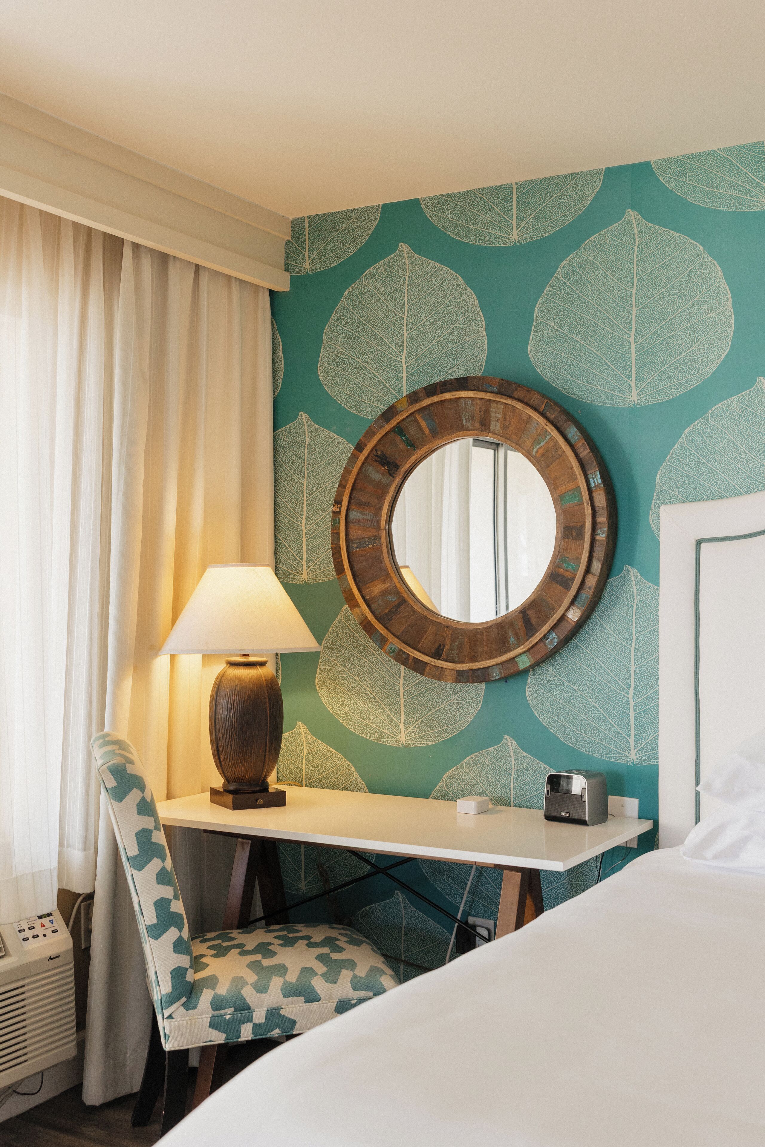 Ein Hotelzimmer mit Bett, Schreibtisch mit Lampe, Stuhl, Spiegel und Wand mit Blattmuster.