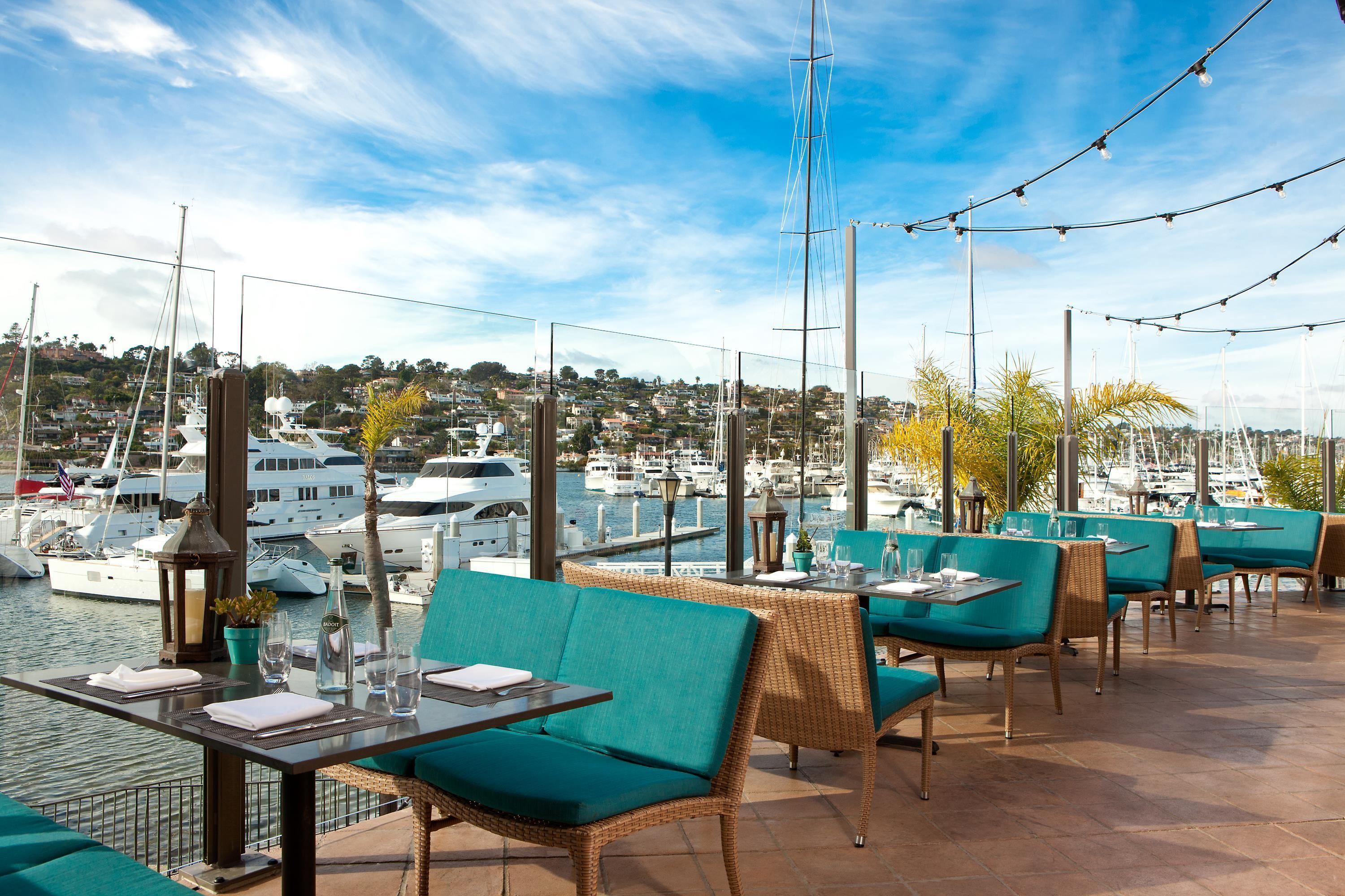 A waterfront restaurant with outdoor seating, overlooking a marina with boats and a cityscape in the background.