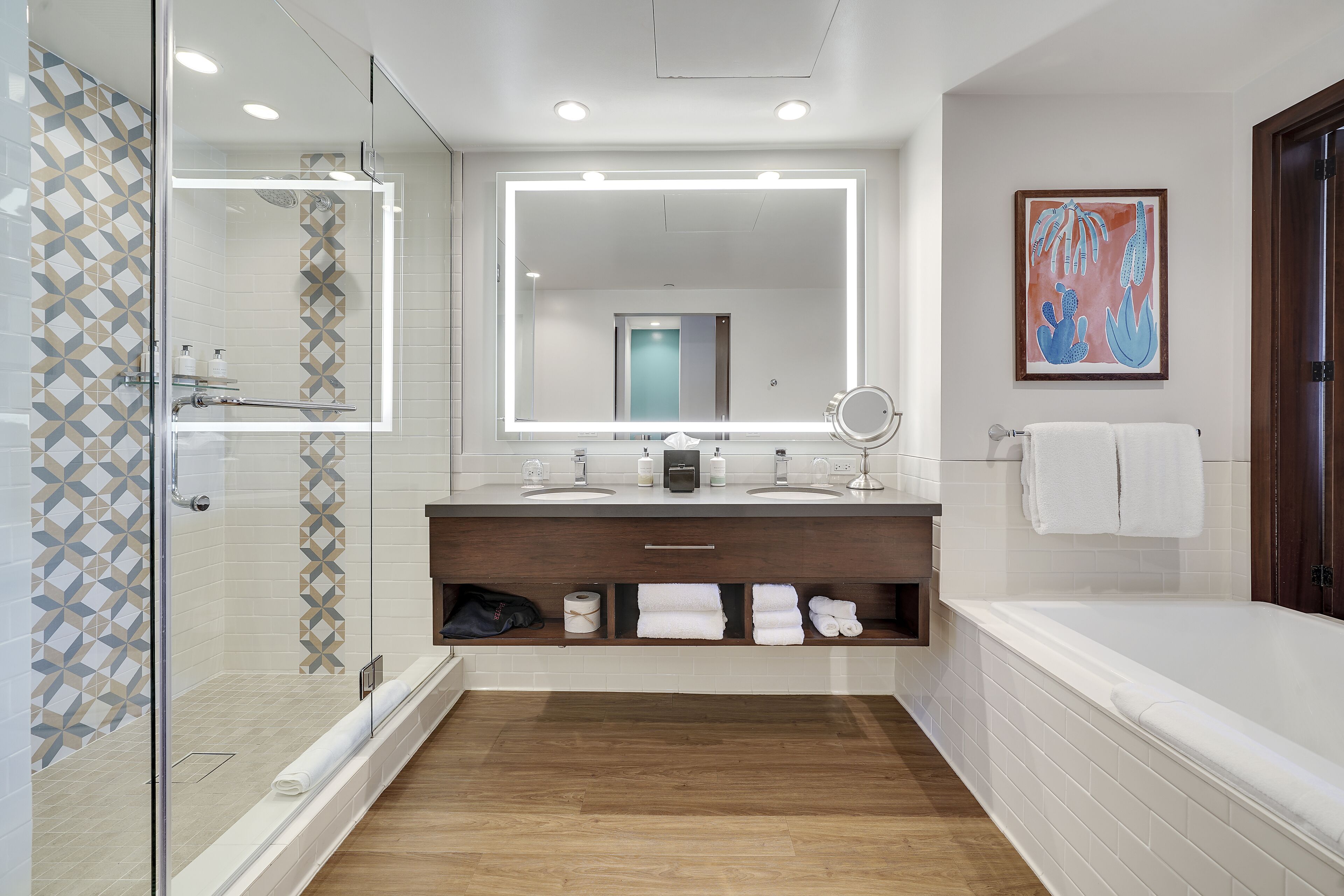 A modern bathroom with a glass-enclosed shower, a large mirror with lights, a bathtub, and a vanity with a towel rack.