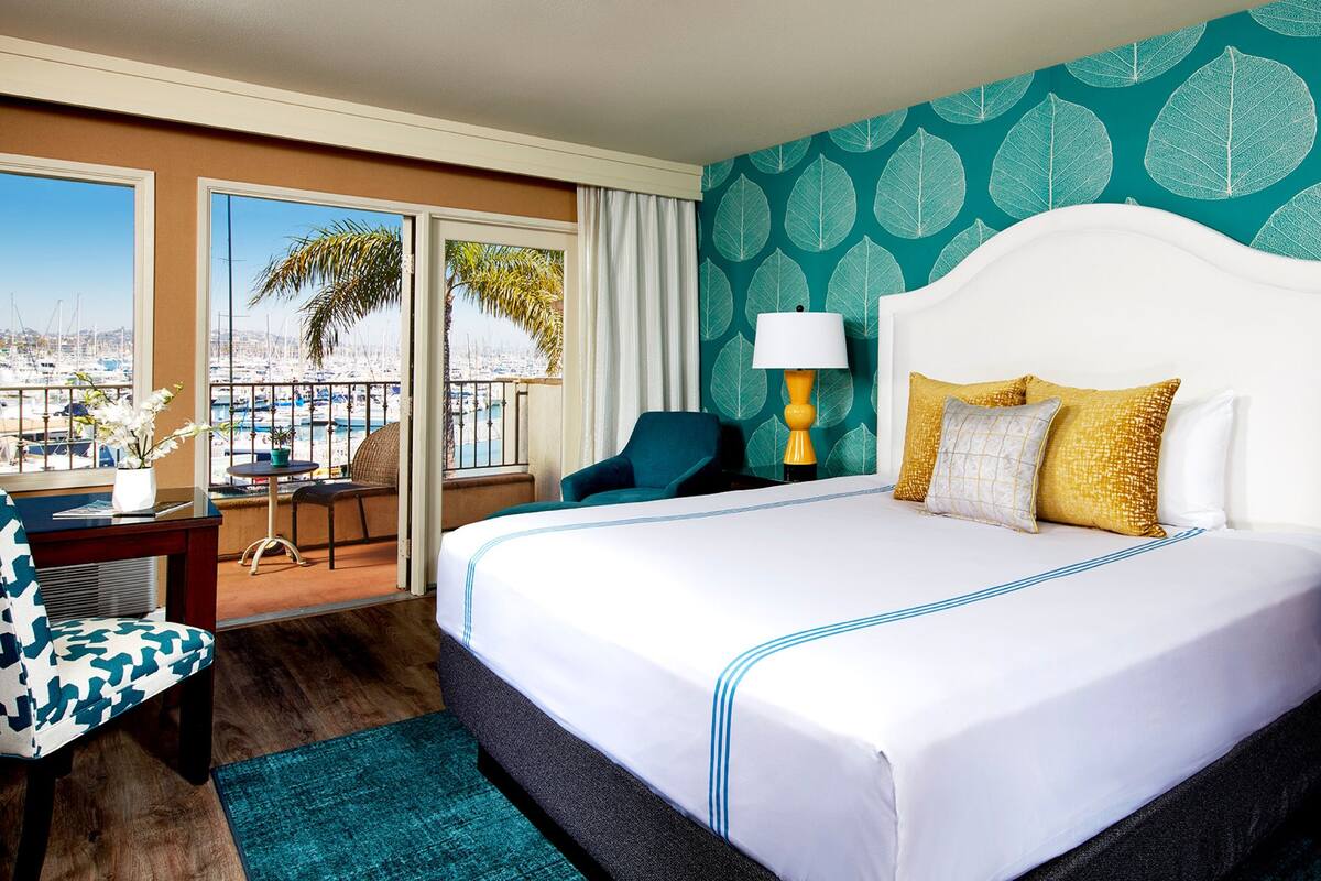 Une chambre d’hôtel avec un grand lit, un bureau avec une chaise, un balcon donnant sur le port de plaisance et un mur décoré de motifs.