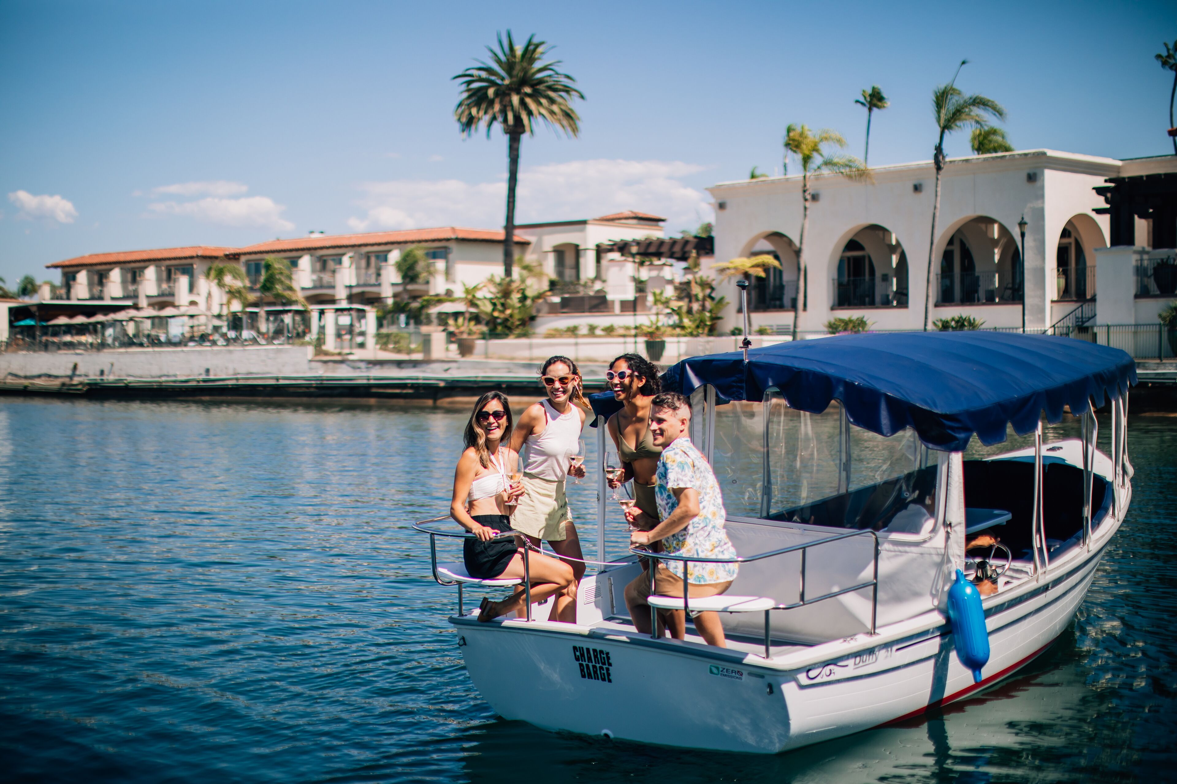 A boat with a blue canopy and a "Charge Range" sign, docked near a waterfront with buildings and palm trees in the background.