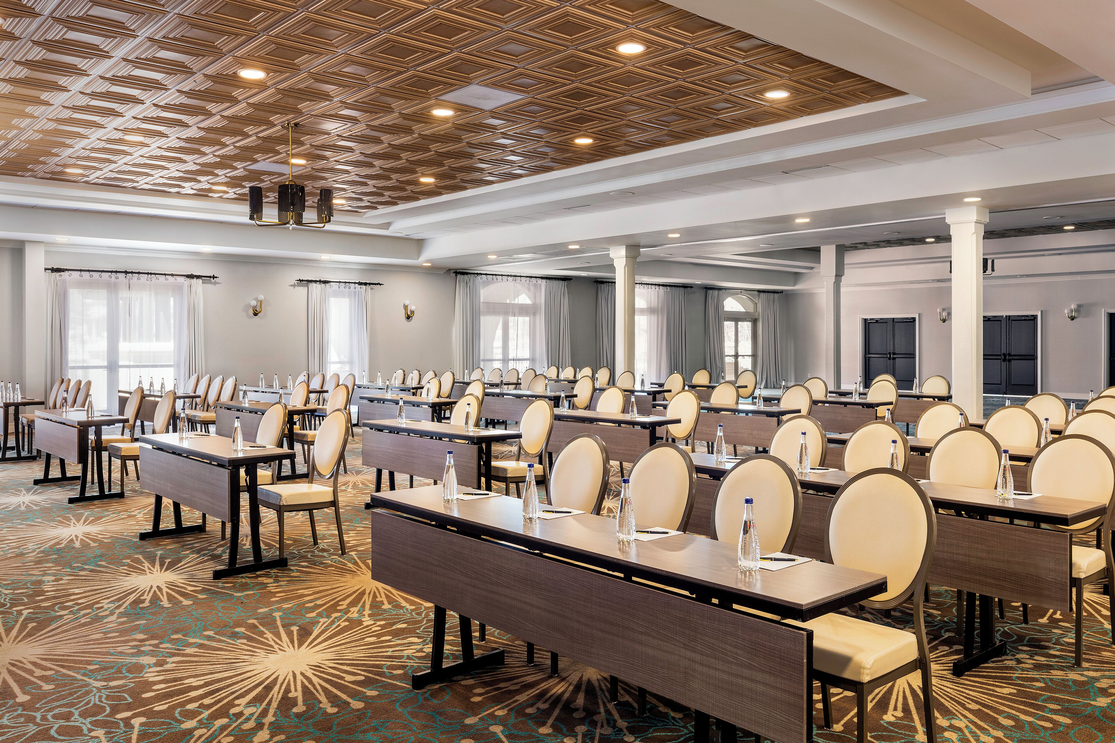 A conference room with rows of tables and chairs, a patterned carpet, and a wooden ceiling with recessed lighting.
