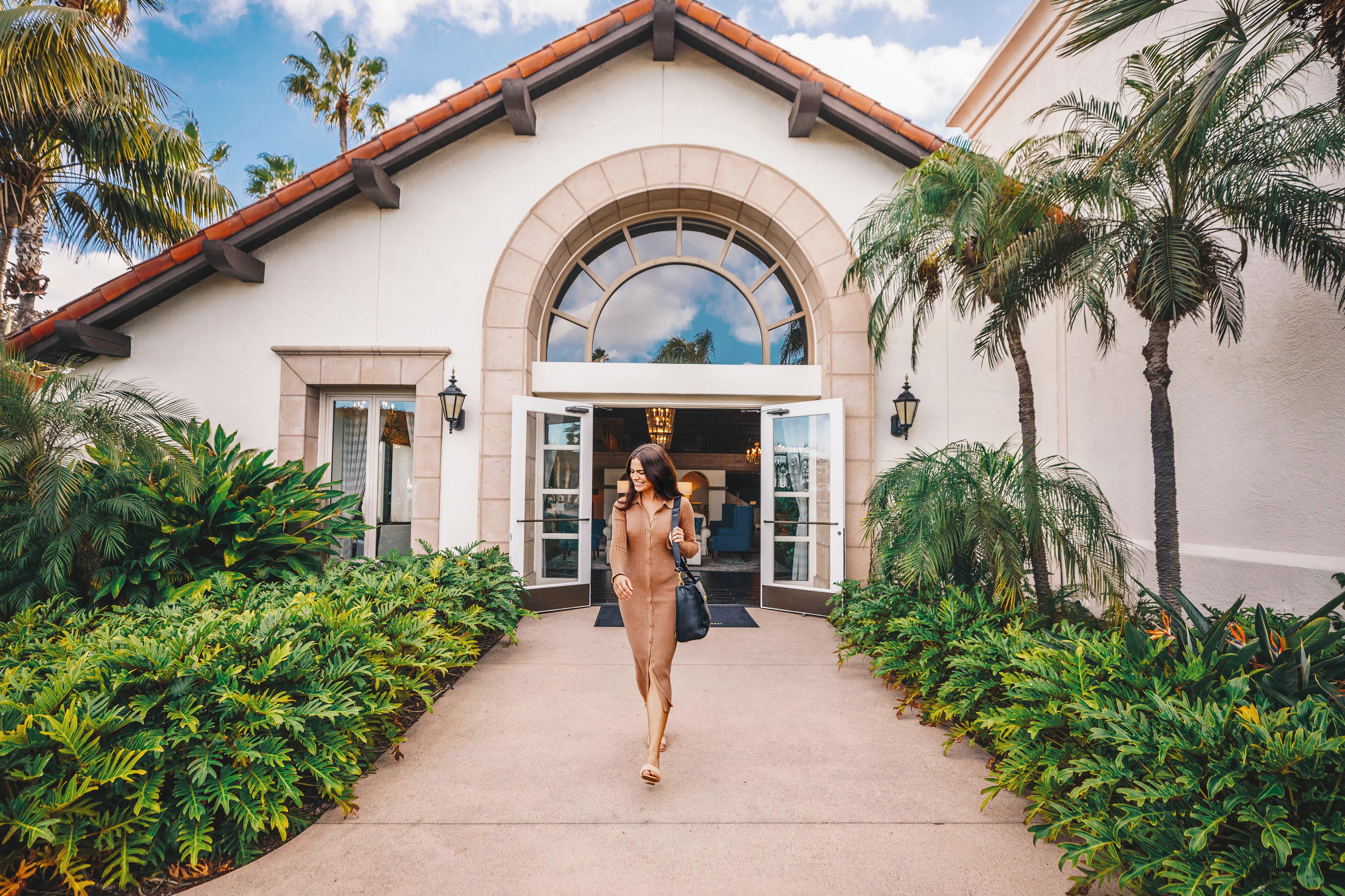 A woman walking towards a building with a large arched entrance, surrounded by lush greenery.