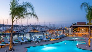 Zona de piscina con sillones de descanso y sombrillas, un bar y vista a barcos al fondo.