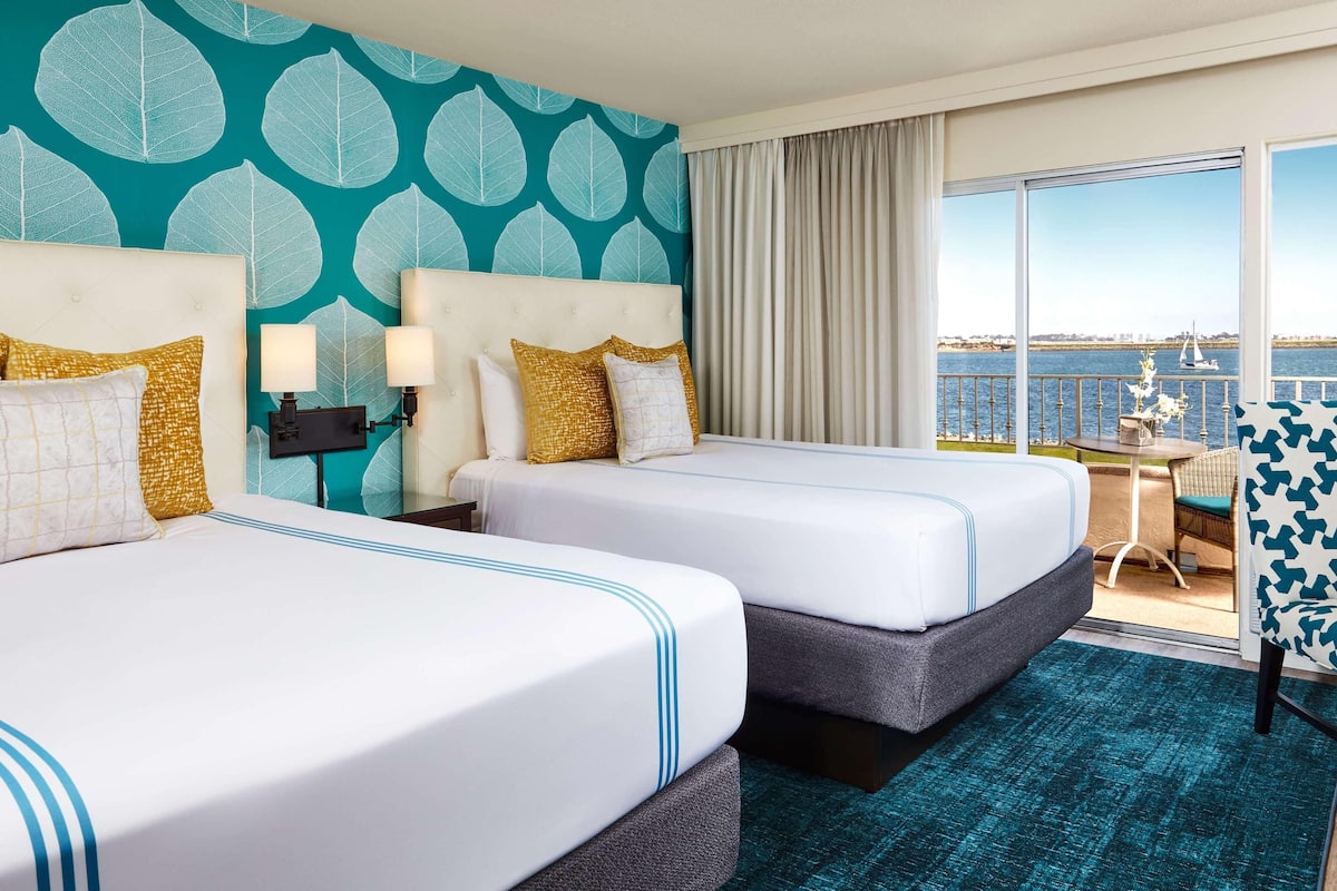 Une chambre d’hôtel avec deux lits, une grande fenêtre donnant sur la mer et un balcon avec une table et une chaise.