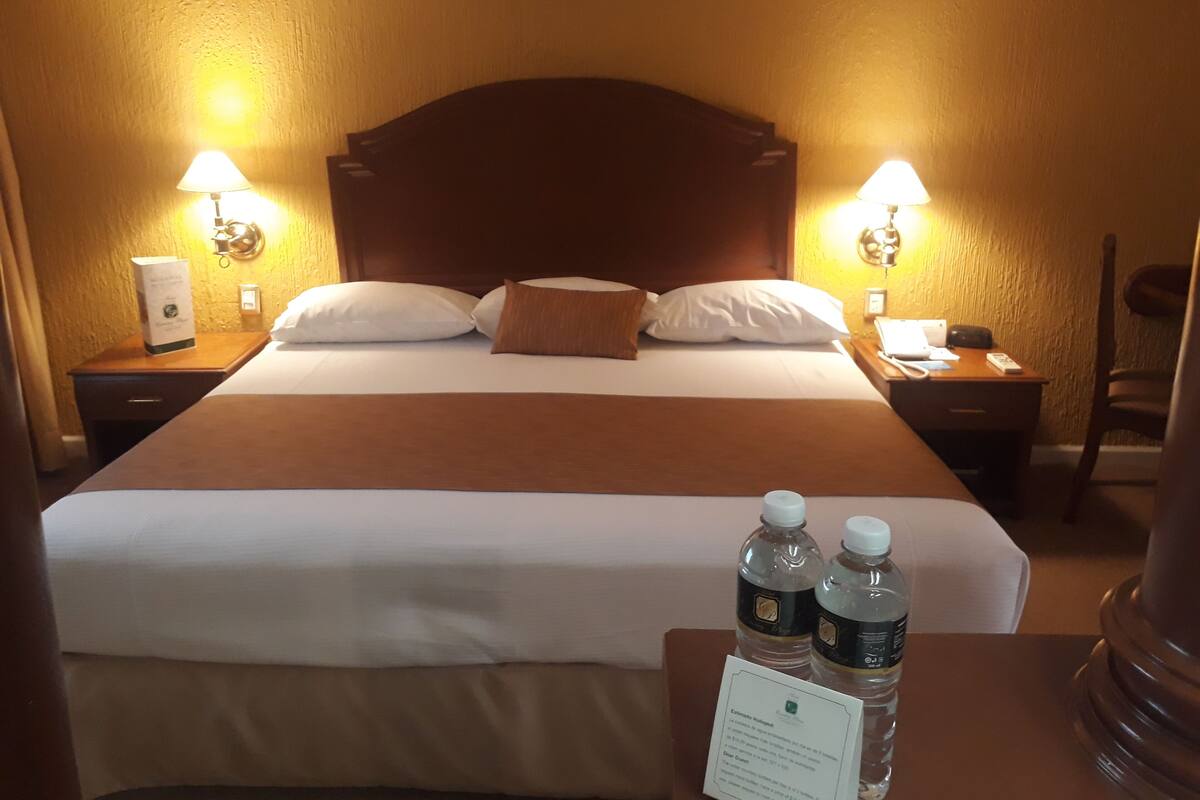 A hotel room with a large bed, two bedside lamps, a nightstand with a phone, and a chair. There are two water bottles on the bed.