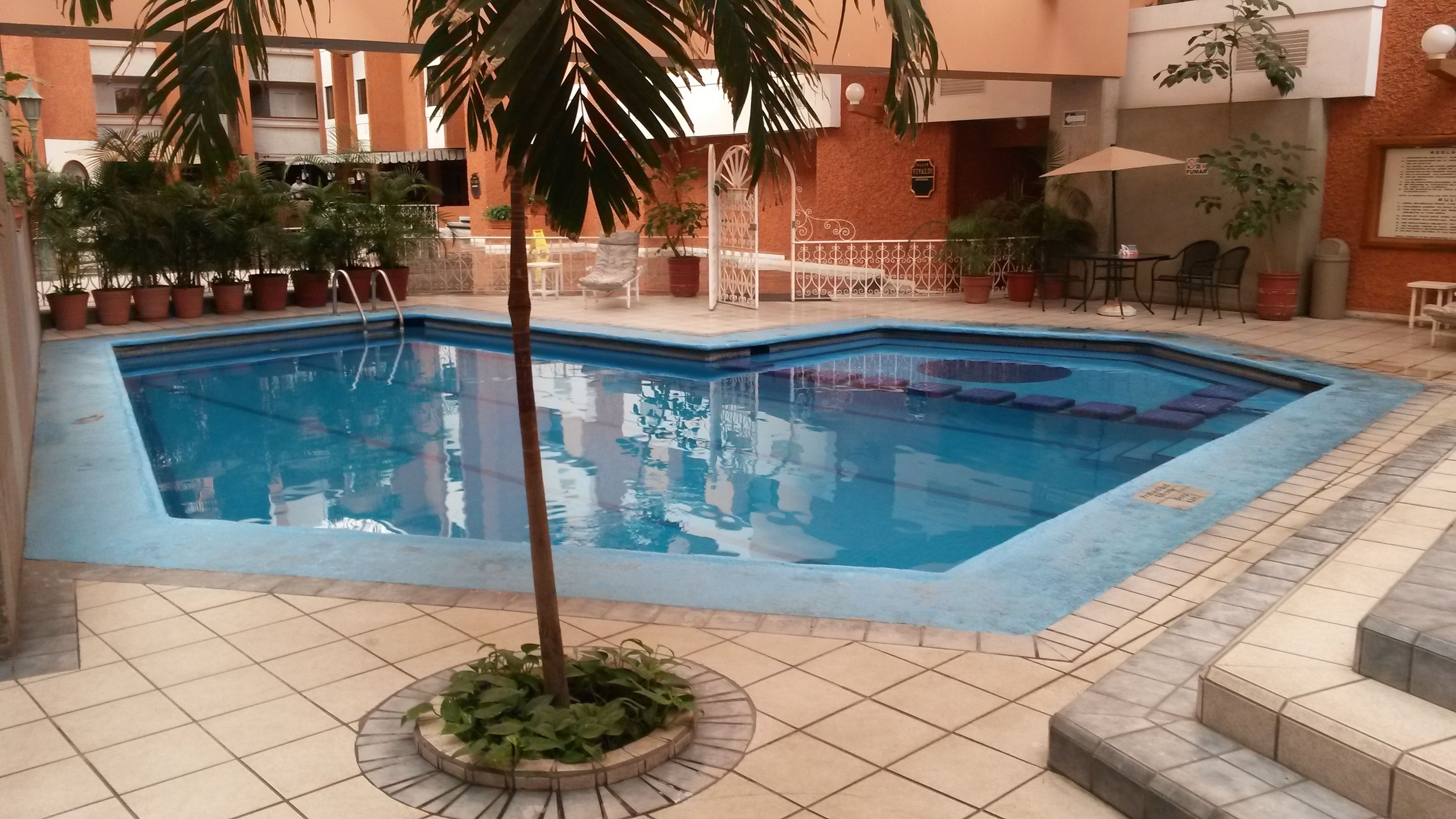 An indoor swimming pool with tiled flooring, surrounded by potted plants and outdoor seating.