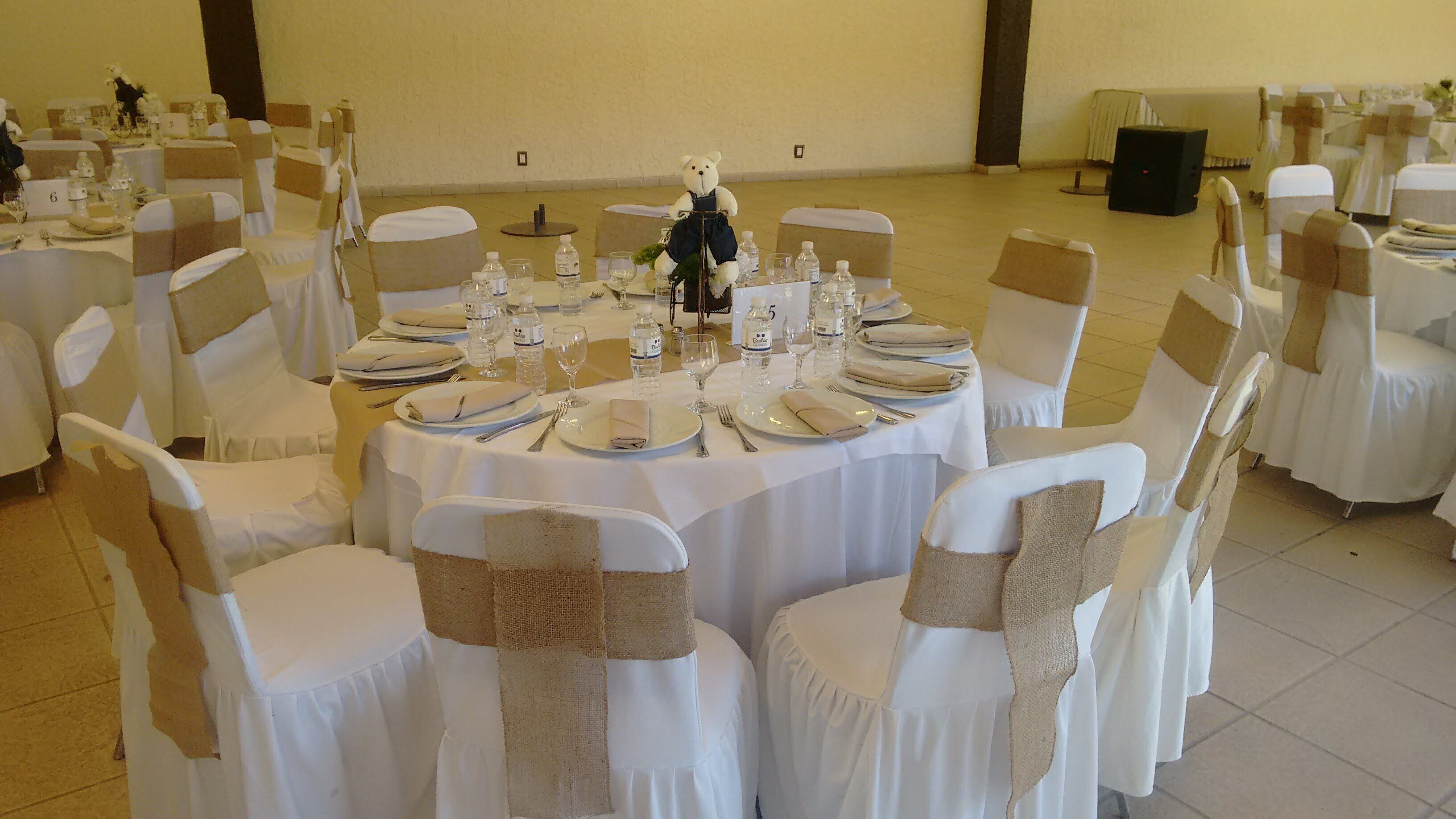 Une salle de banquet avec des tables rondes dressées pour un événement formel, chacune étant recouverte d’une nappe blanche et agrémentée de couverts en argent et de bouteilles d’eau.