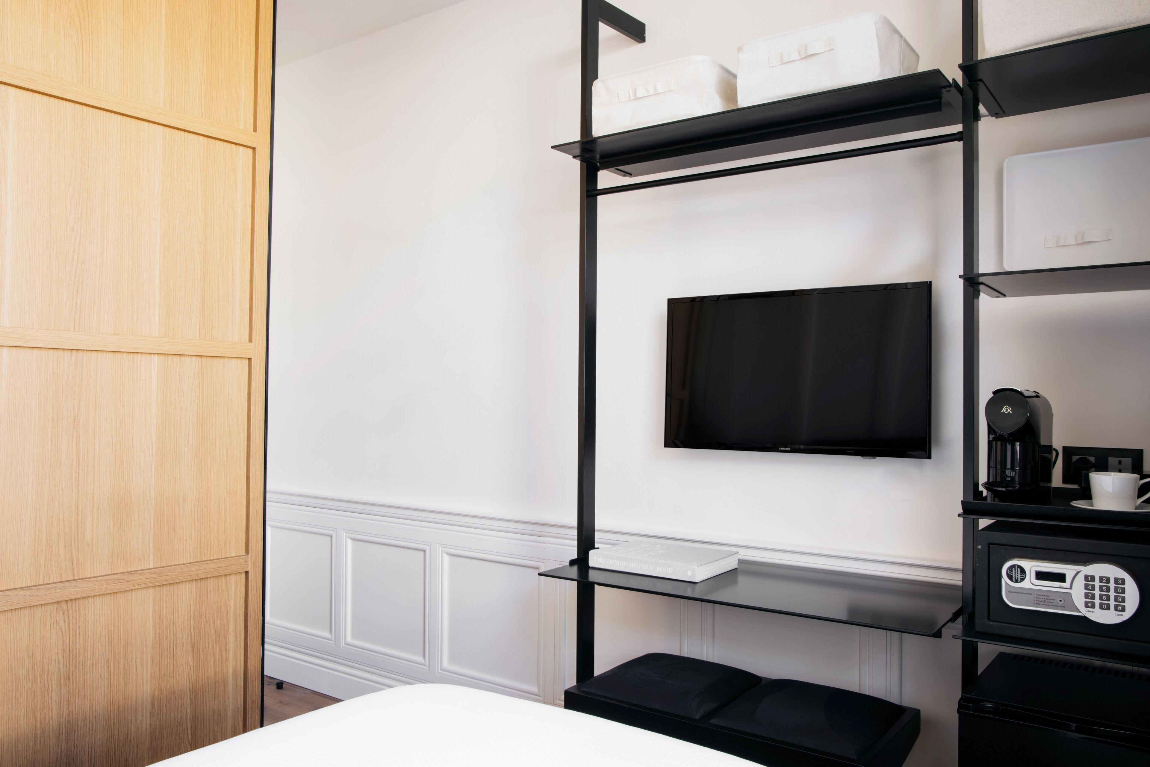 A hotel room with a wooden wardrobe, a black TV mounted on the wall, and a black shelf unit with a coffee machine and a digital device.