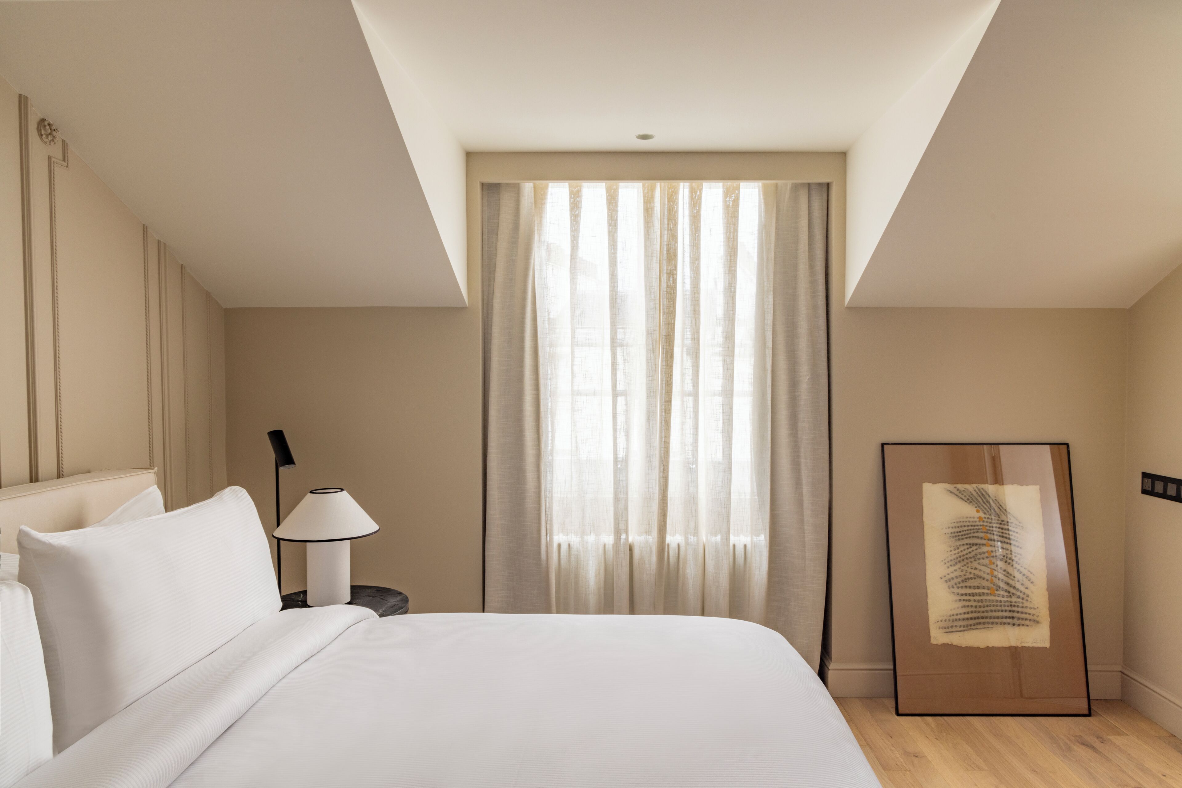 A hotel room with a large bed, a nightstand with a lamp, a window with sheer curtains, and a framed artwork on the wall.