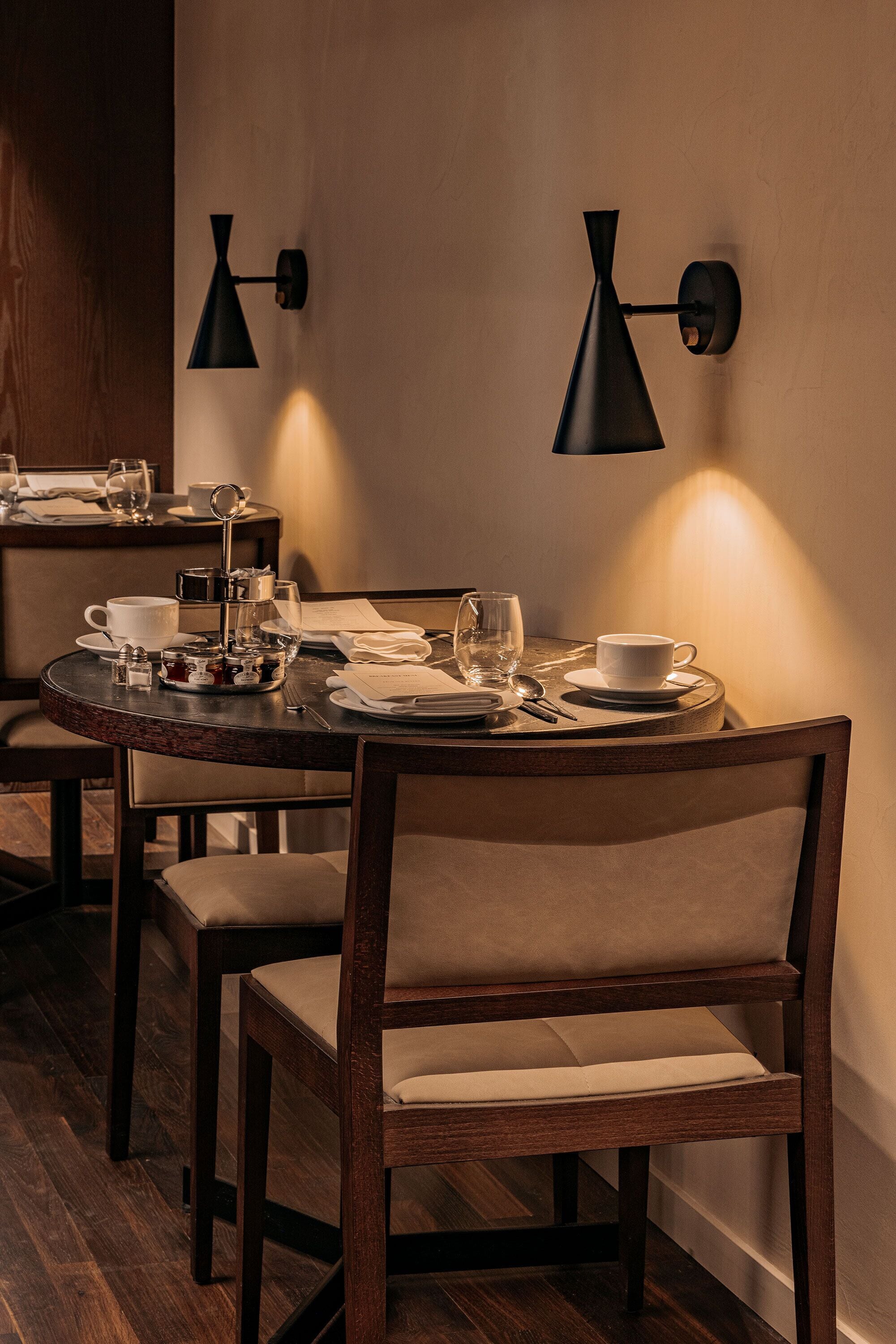 Un comedor con una mesa redonda dispuesta para dos personas, que incluye un candelabro, platos y copas. Dos lámparas de pared proporcionan iluminación.
