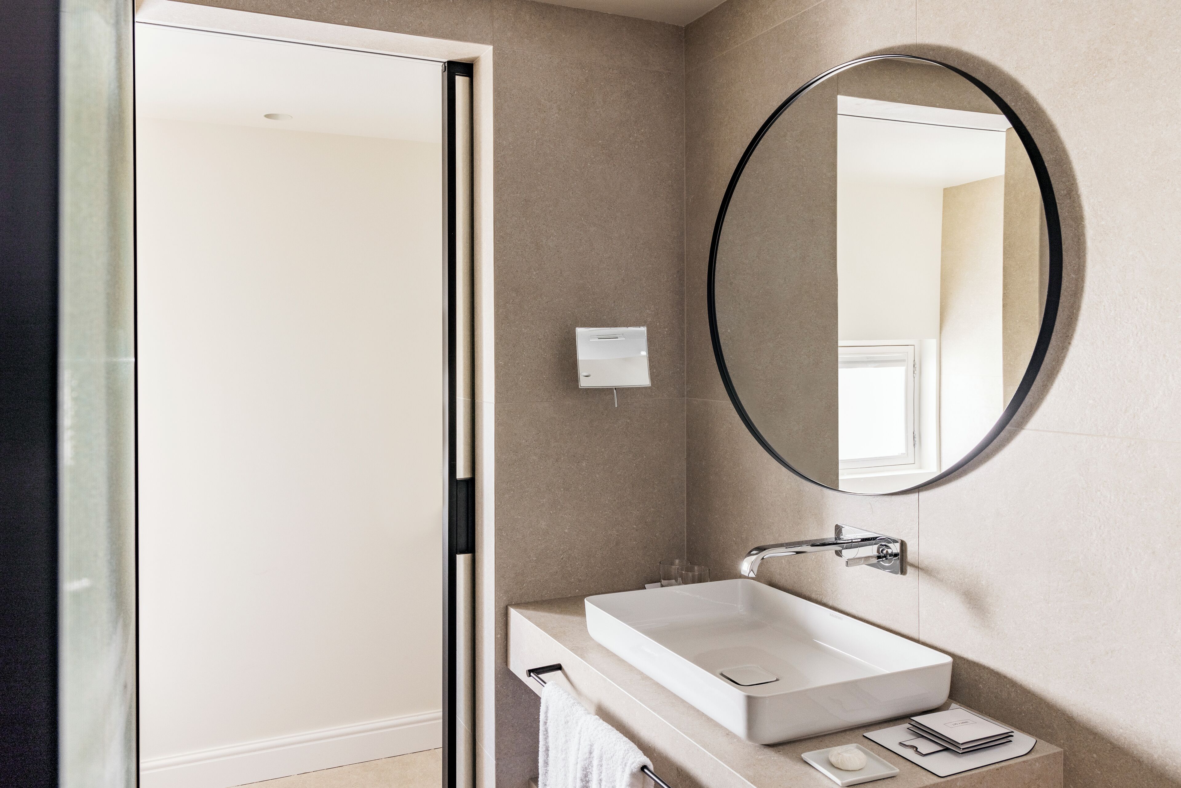 Un baño moderno con un lavamanos blanco, un espejo redondo y un perchero para toallas.