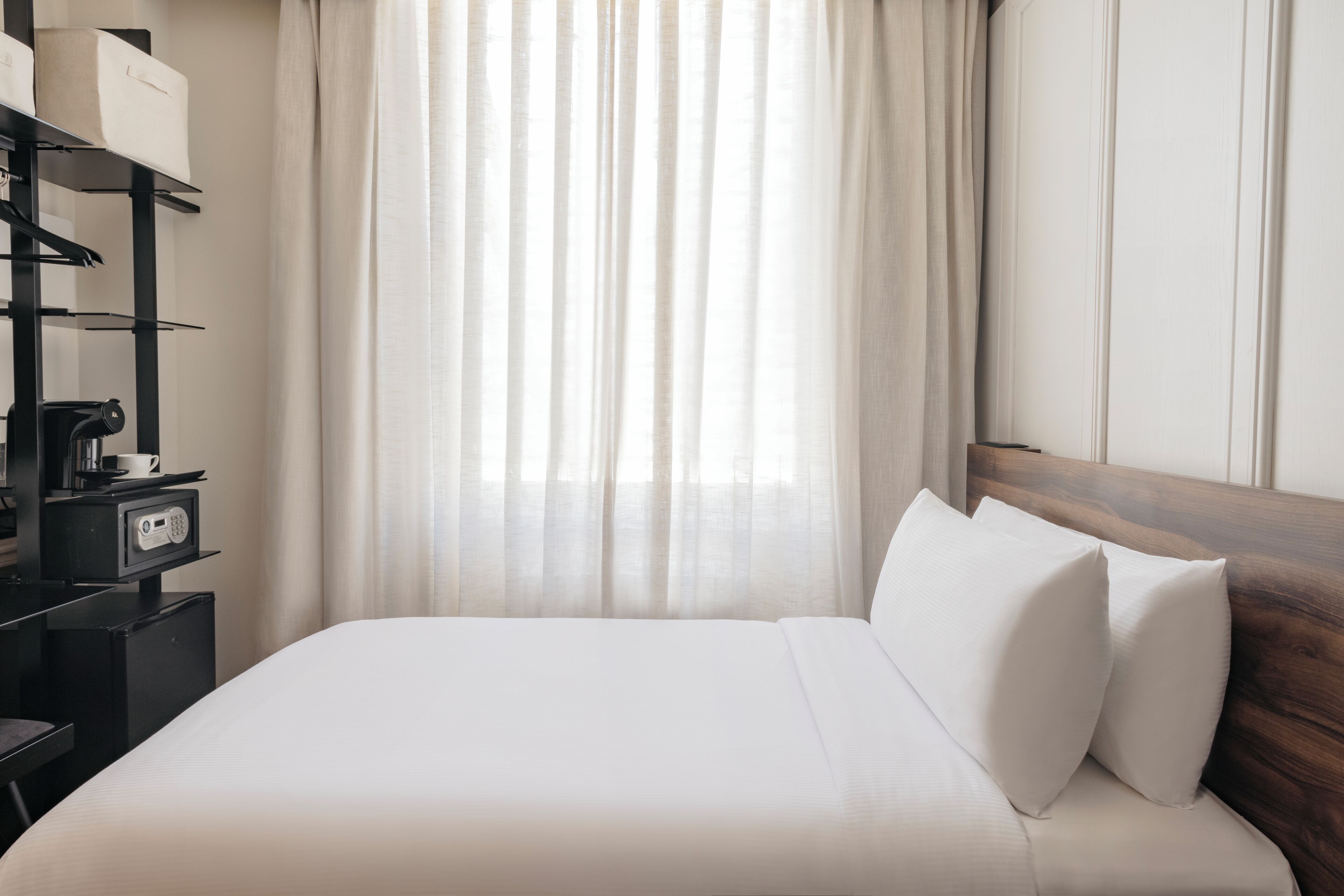 A hotel room with a neatly made bed, a bedside table with a coffee maker, and a wardrobe with hanging clothes.