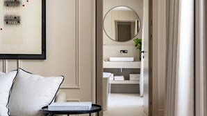 Una habitación de hotel moderna con una paleta de colores neutros, que incluye un sofá, una mesita redonda pequeña, una obra de arte enmarcada y vistas al baño con un espejo y lavamanos.