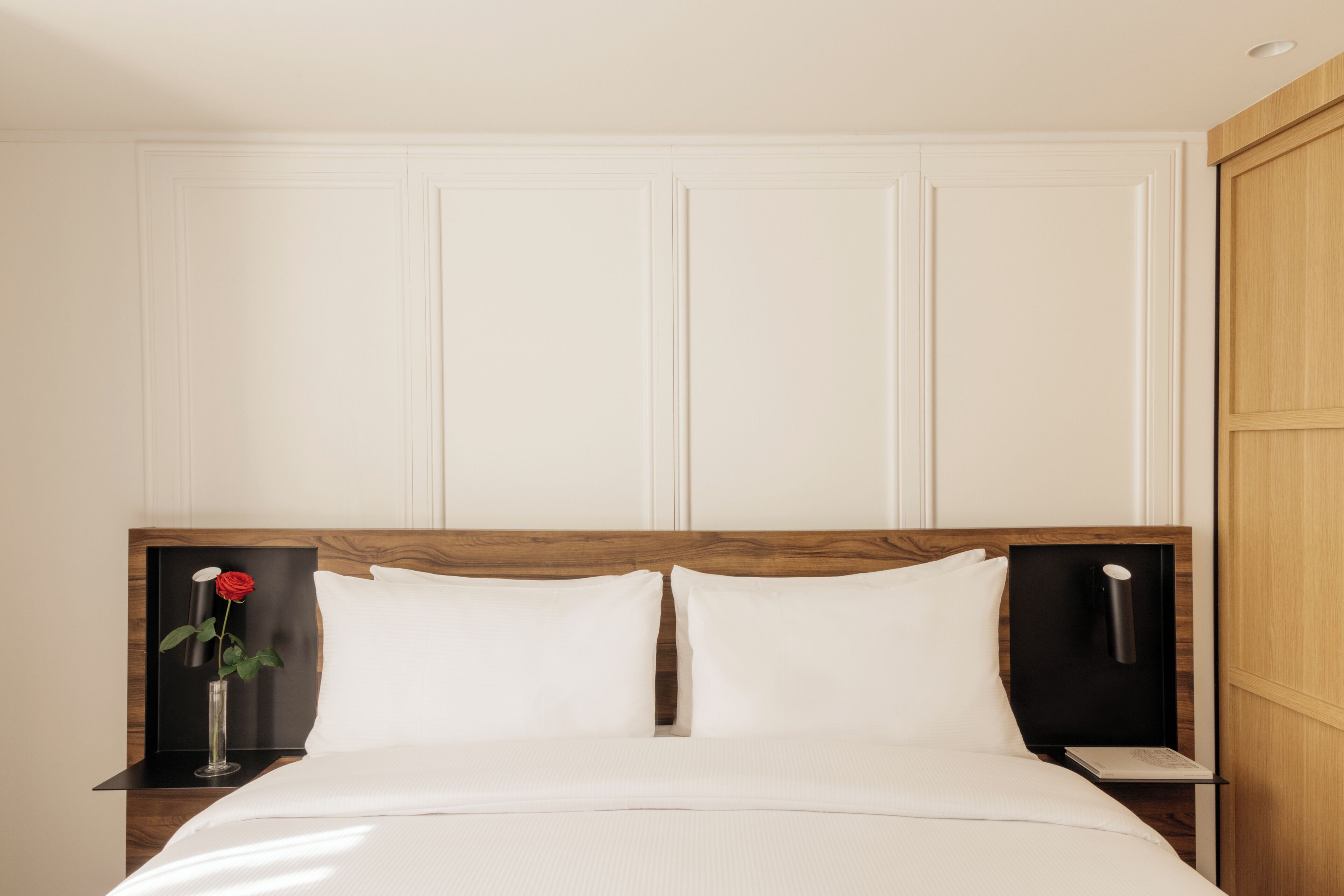 Una cama bien hecha con sábanas blancas, cabecera de madera y mesita de noche con un florero de rosa roja.