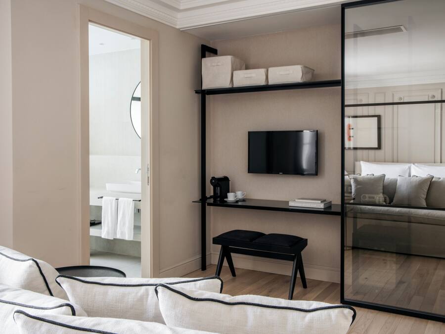 Ein modernes Hotelzimmer mit einer Couch, einem schwarzen Couchtisch, einem Flachbildfernseher, der an einem schwarzen Regal befestigt ist, und einer Glastrennwand, die den Wohnbereich vom Schlafzimmer abgrenzt.