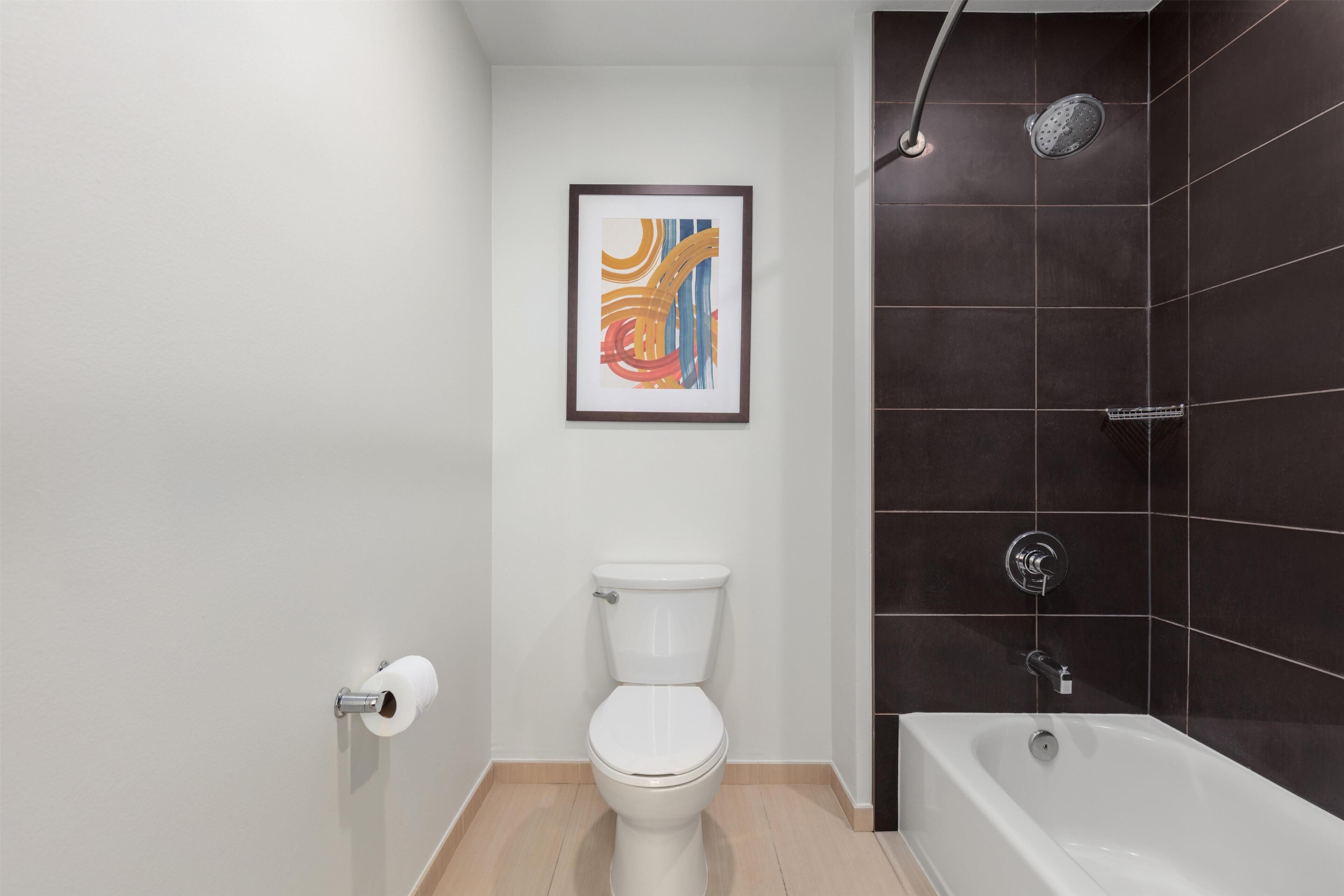 A modern bathroom with a white toilet, a framed abstract painting, and a dark tiled shower area.