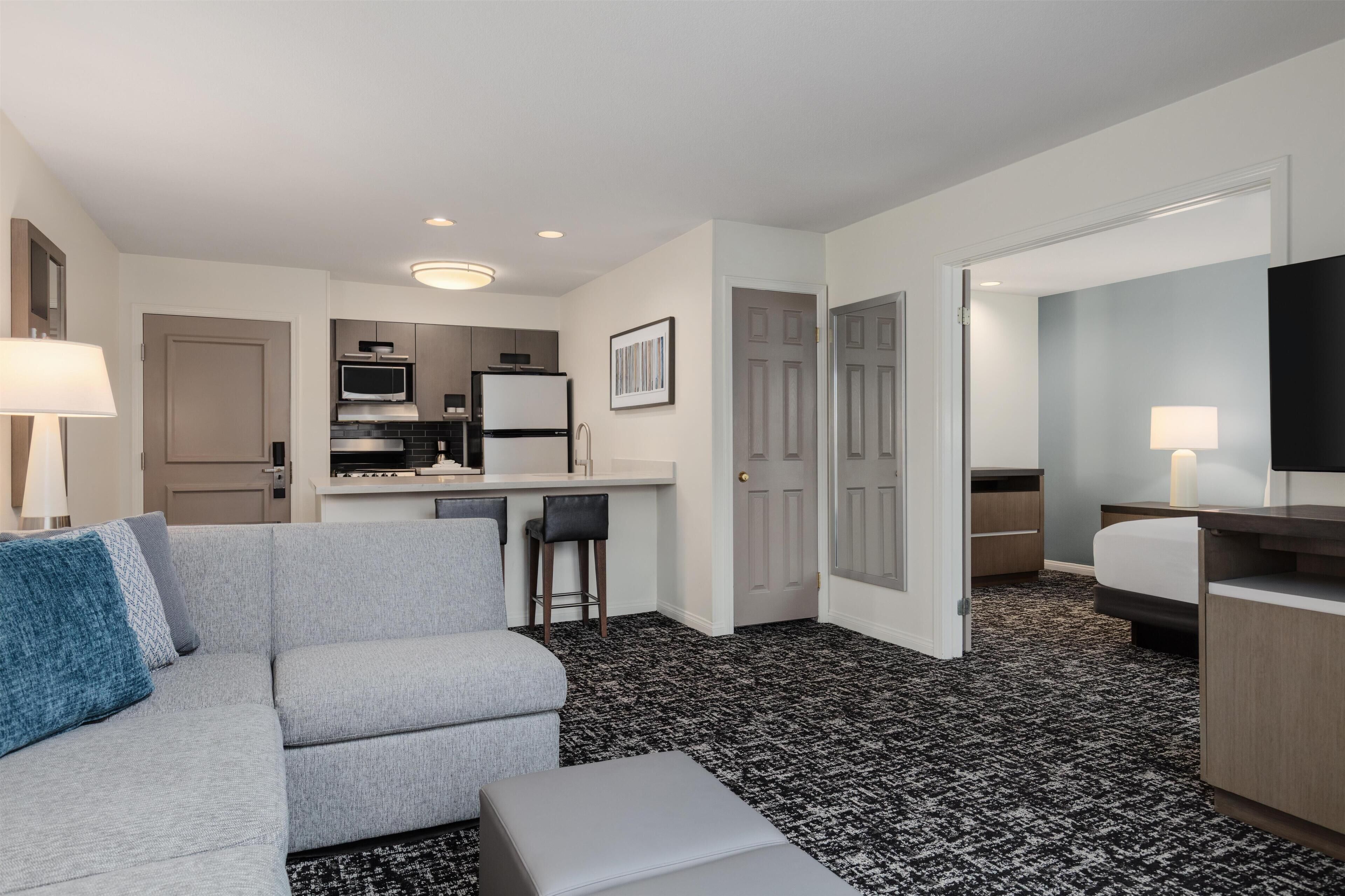 A modern hotel room with a grey sofa, a small dining area, a kitchenette, a TV, and a patterned carpet.