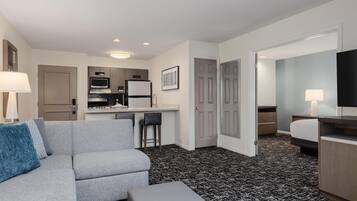 A modern hotel room with a grey sofa, a small dining area, a kitchenette, a TV, and a patterned carpet.