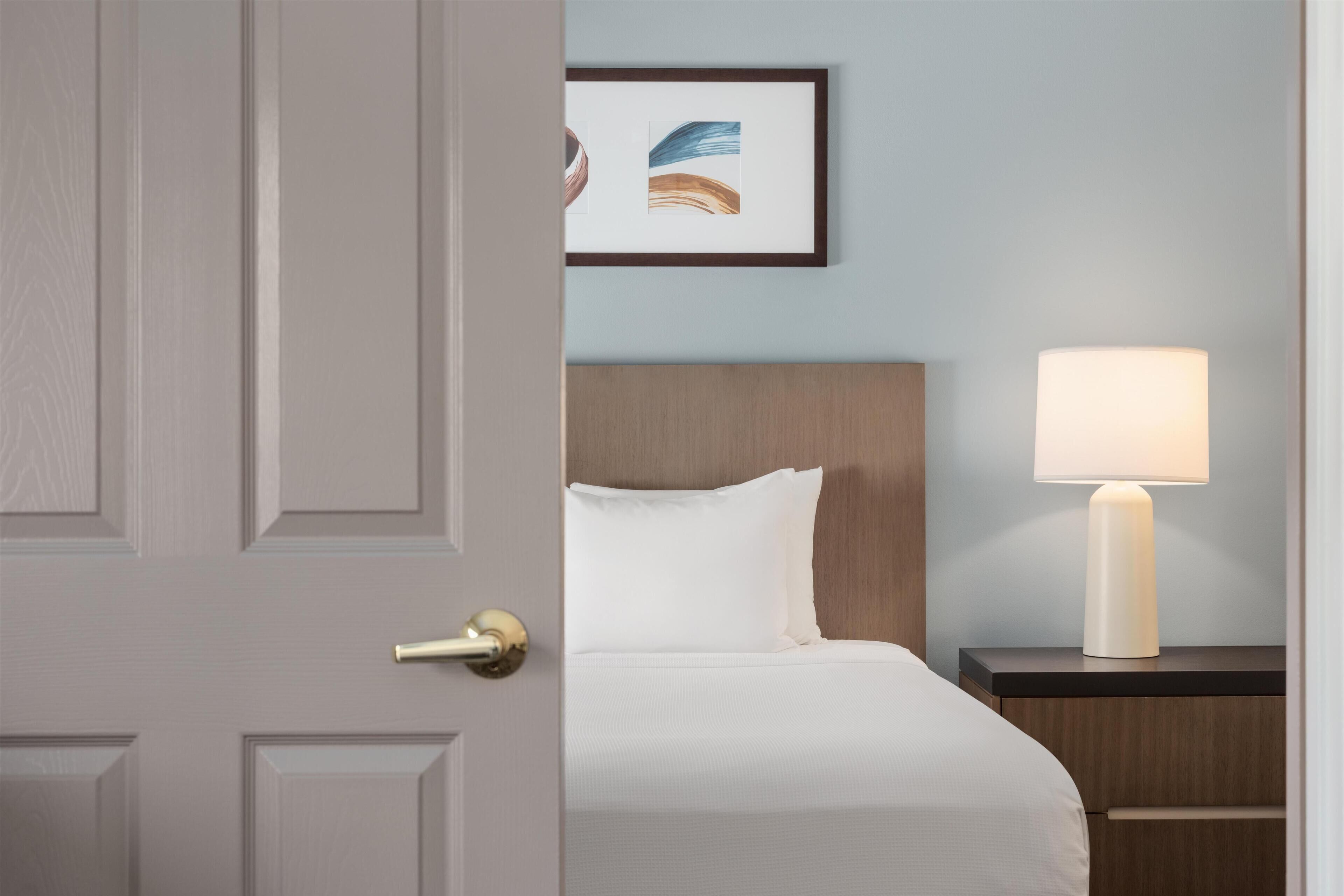 Quarto de hotel com uma cama, mesa de cabeceira com abajur e um quadro na parede.