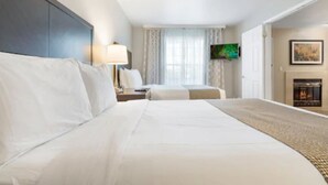 A hotel room with a large bed, a TV mounted on the wall, a fireplace, and a window with curtains.