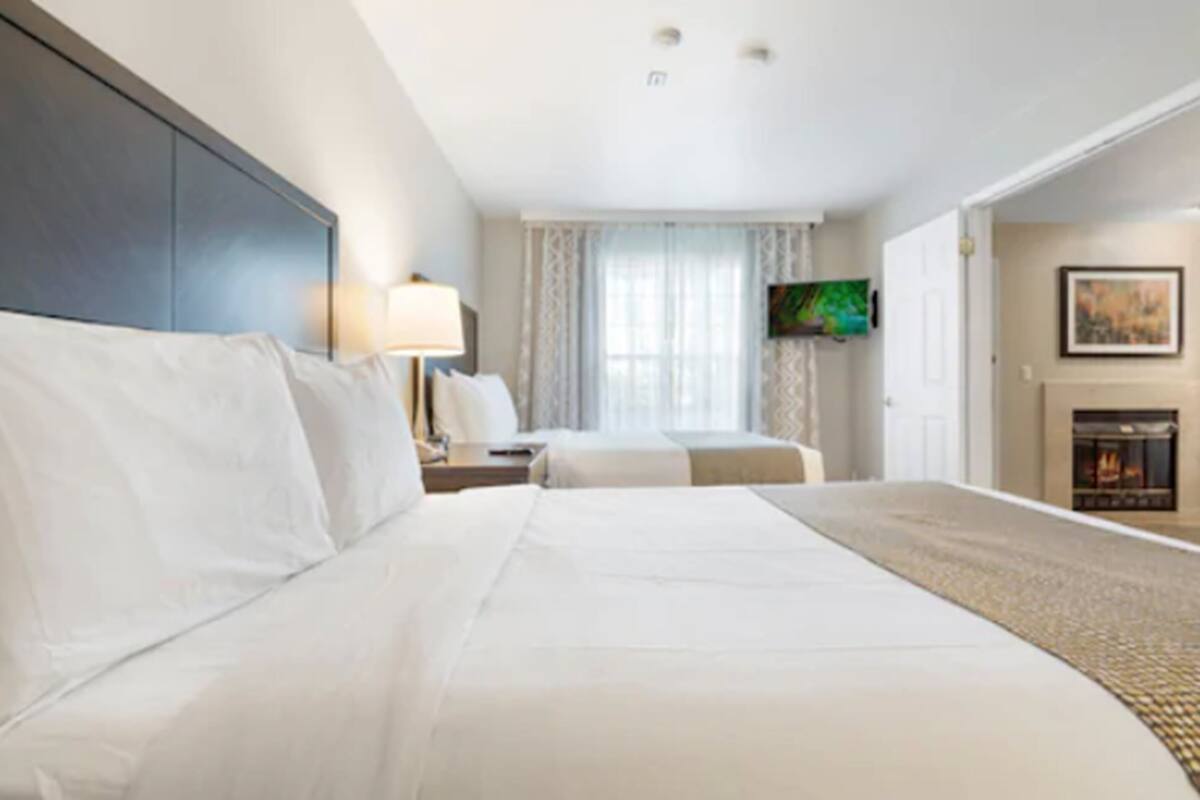 A hotel room with a large bed, a TV mounted on the wall, a fireplace, and a window with curtains.