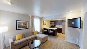 A hotel room with a sofa, a small coffee table, a flat-screen TV, a kitchenette with a microwave, refrigerator, and a dining area with chairs.