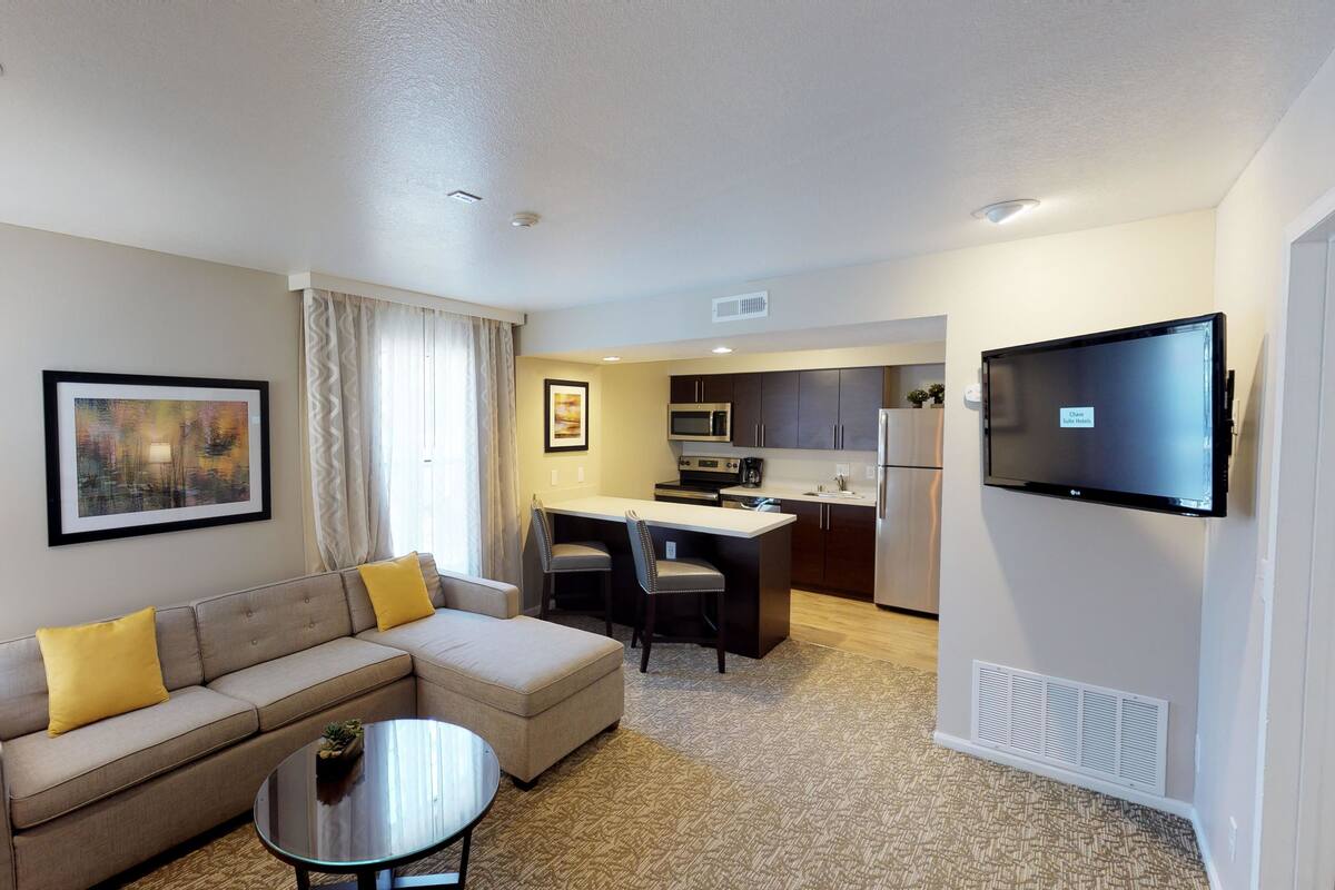 A hotel room with a sofa, a small coffee table, a flat-screen TV, a kitchenette with a microwave, refrigerator, and a dining area with chairs.