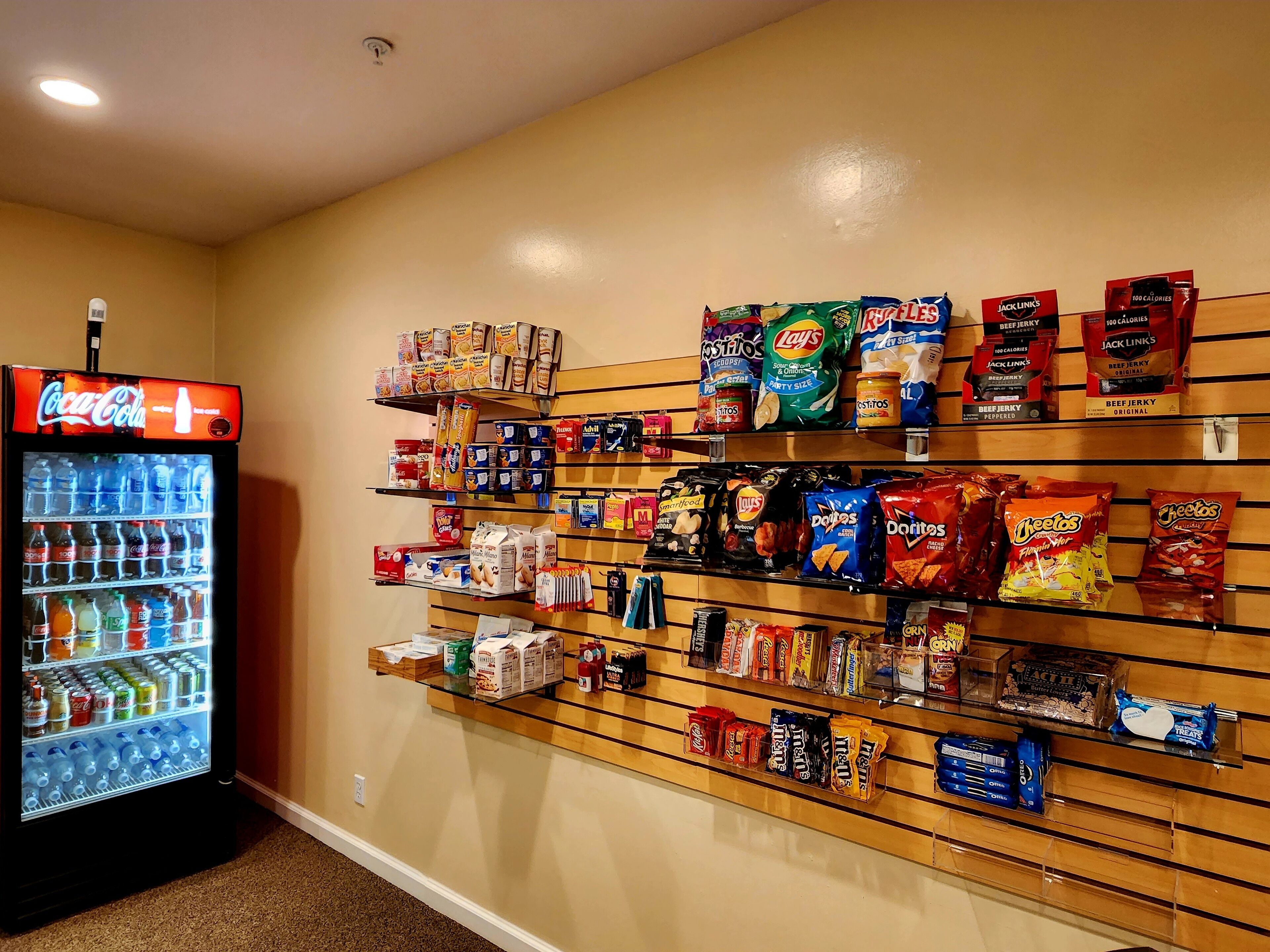 Een snackwand met verschillende merken en soorten chips, een Coca-Cola koelkast, en een plank met flessen water.