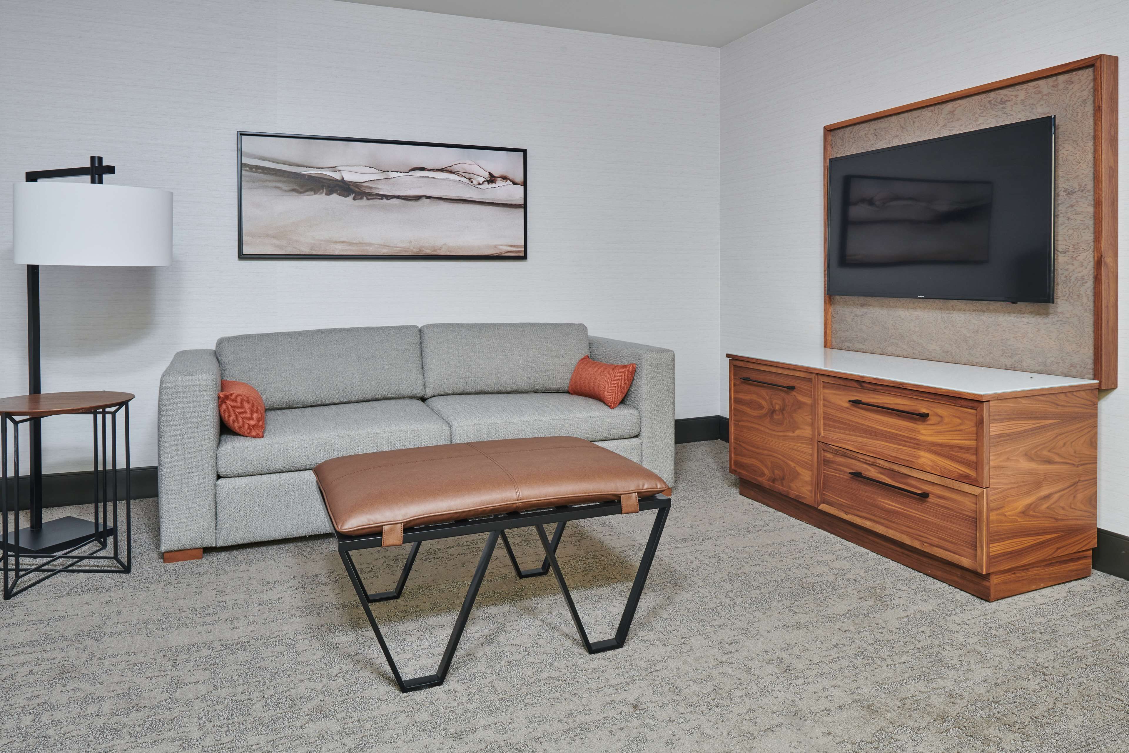 Una sala de estar moderna con un sofá gris, un banco de cuero marrón, un mueble de madera, un televisor y una lámpara de pie.