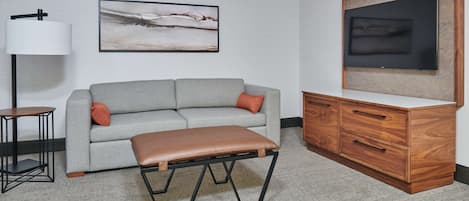 Una sala de estar moderna con un sofá gris, un banco de cuero marrón, un mueble de madera, un televisor y una lámpara de pie.