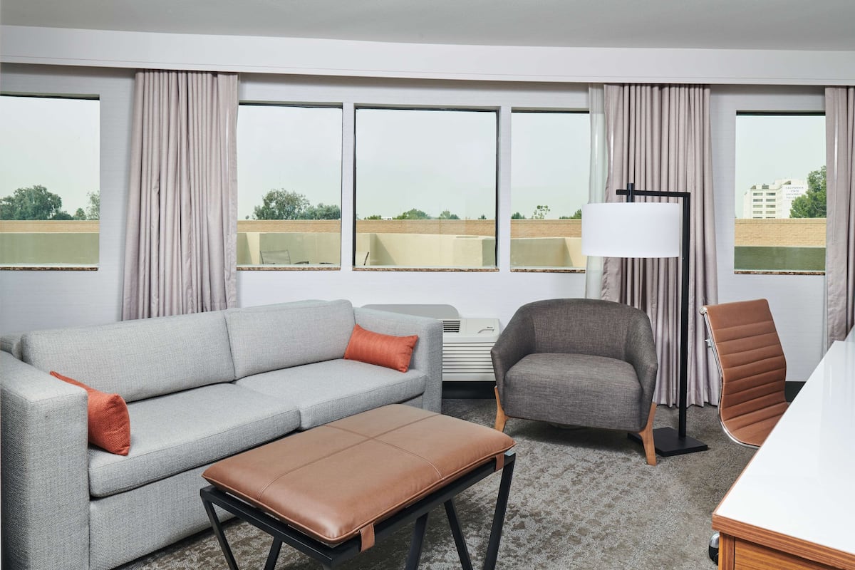 A hotel room with a grey sofa, a brown ottoman, a grey armchair, a standing lamp, and a view of the outside through large windows.