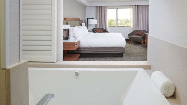 A hotel room with a large bed, a desk with a lamp, a chair, a window with curtains, and a bathtub. - DoubleTree by Hilton Fullerton (Fullerton)