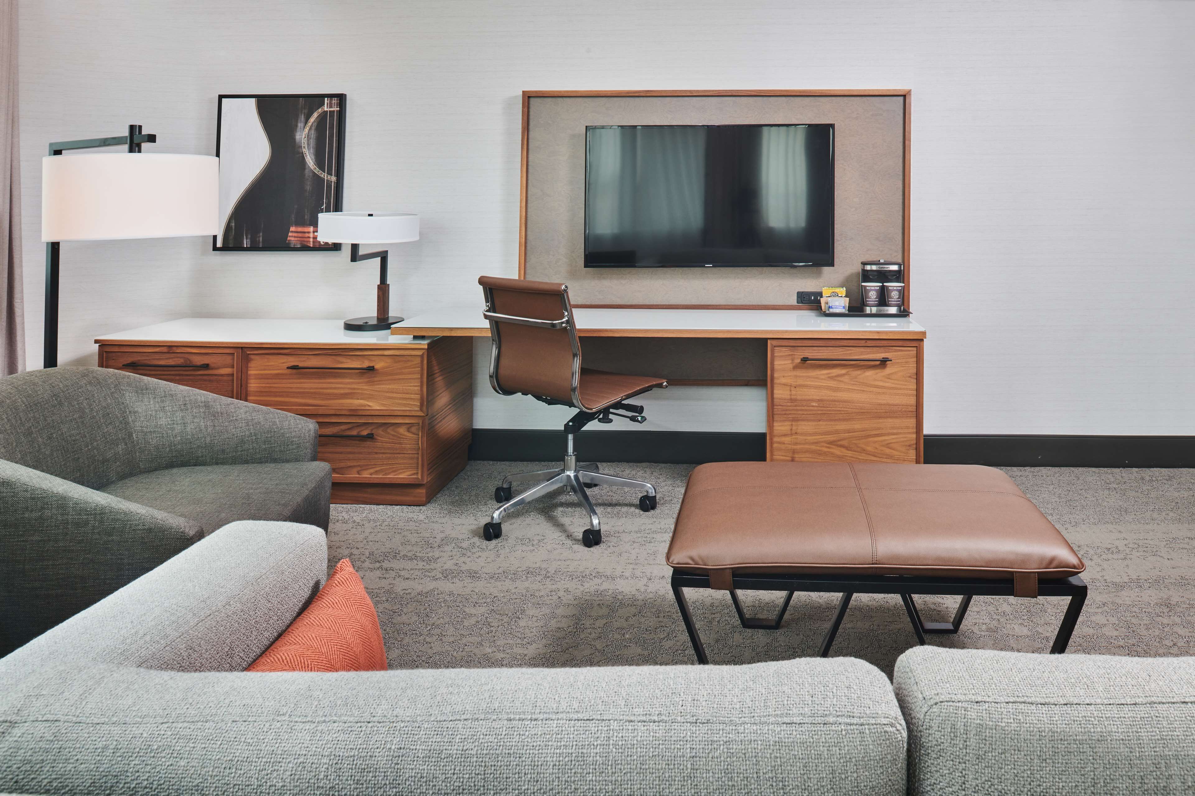 A hotel room with a grey sofa, a brown leather ottoman, a wooden desk with a flat-screen TV, a lamp, and a coffee maker.