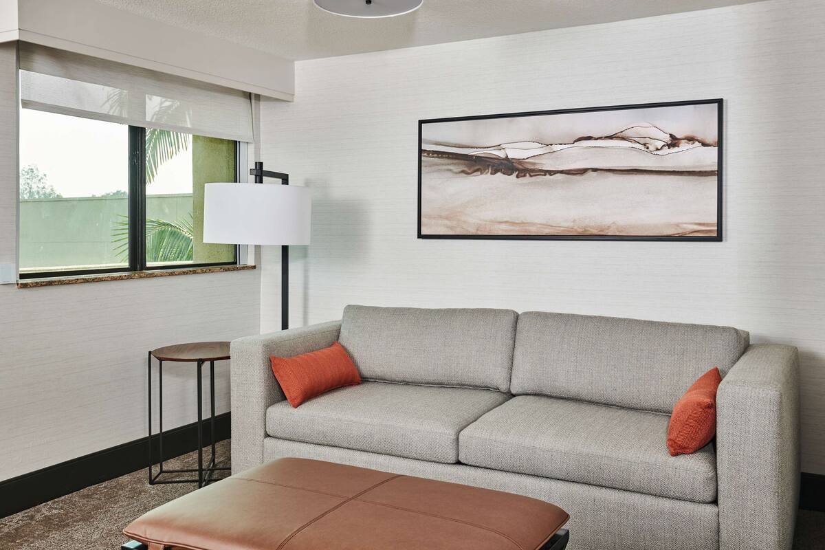 A modern living room with a grey sofa, two orange cushions, a small round table, and a floor lamp. A framed abstract painting is on the wall.