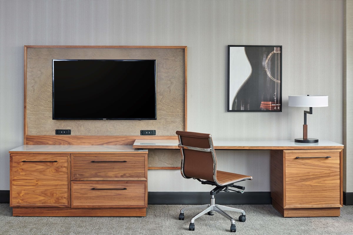 A hotel room with a wooden desk, a flat-screen TV, a modern chair, a lamp, and a framed picture on the wall.