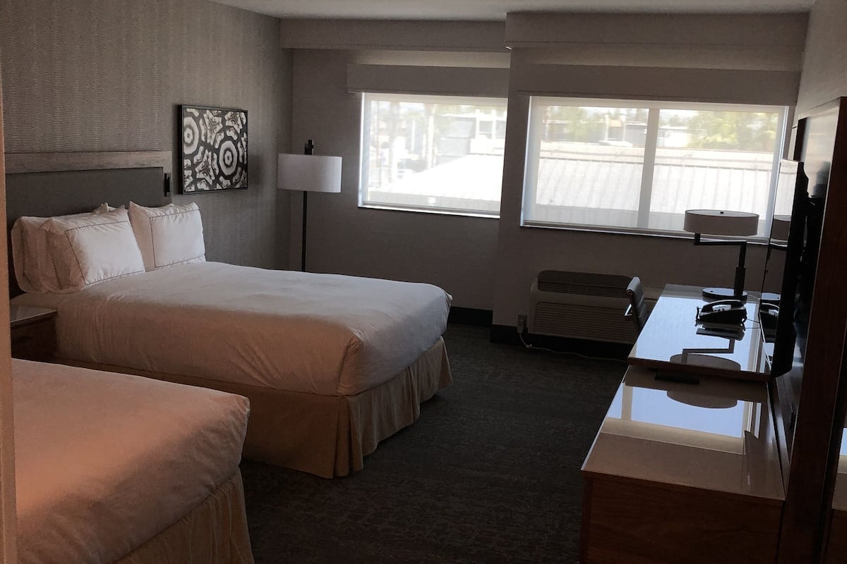 A hotel room with two beds, a desk, a lamp, and a large window.