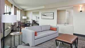A hotel room with a grey sofa, a small table, a brown ottoman, a desk with a chair, and a large window. - DoubleTree by Hilton Fullerton (Fullerton)