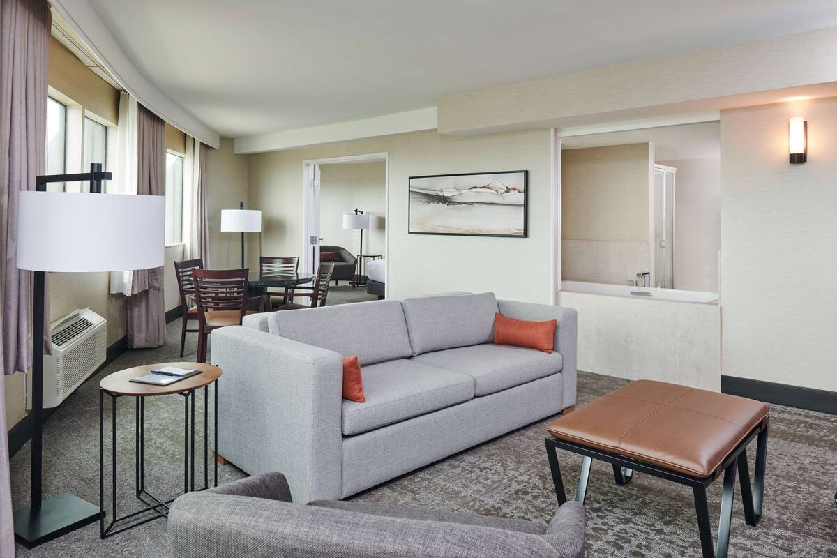 A hotel room with a grey sofa, a small table, a brown ottoman, a desk with a chair, and a large window.