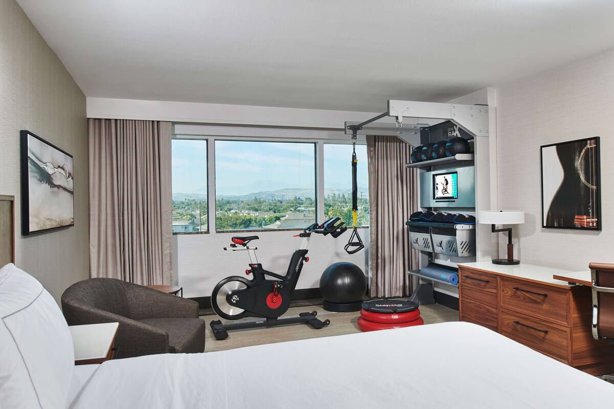 A hotel room with a bed, exercise bike, exercise ball, and a desk with a lamp.