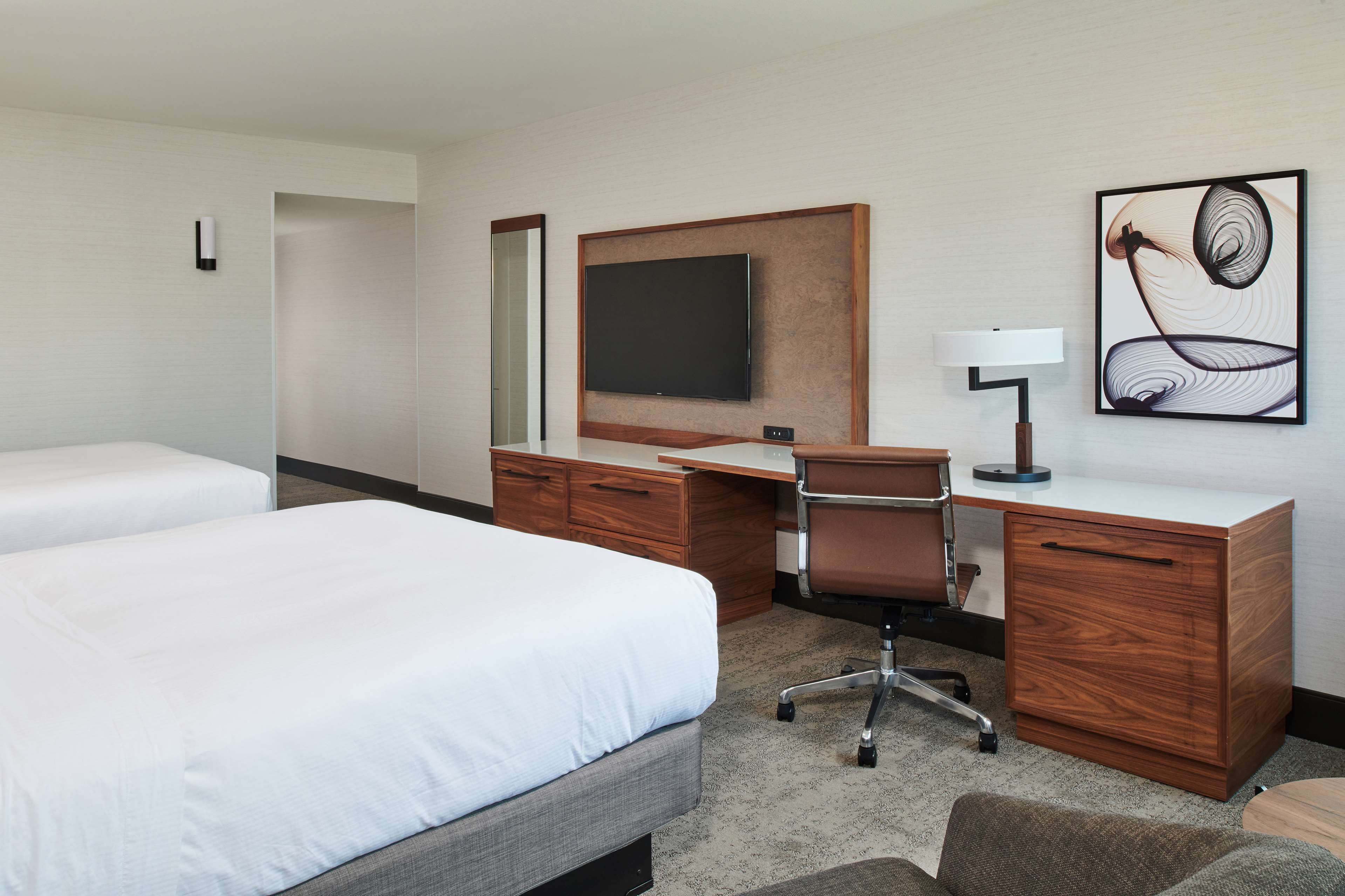 Una habitación de hotel con cama, escritorio, silla, televisor, lámpara y una obra de arte en la pared.