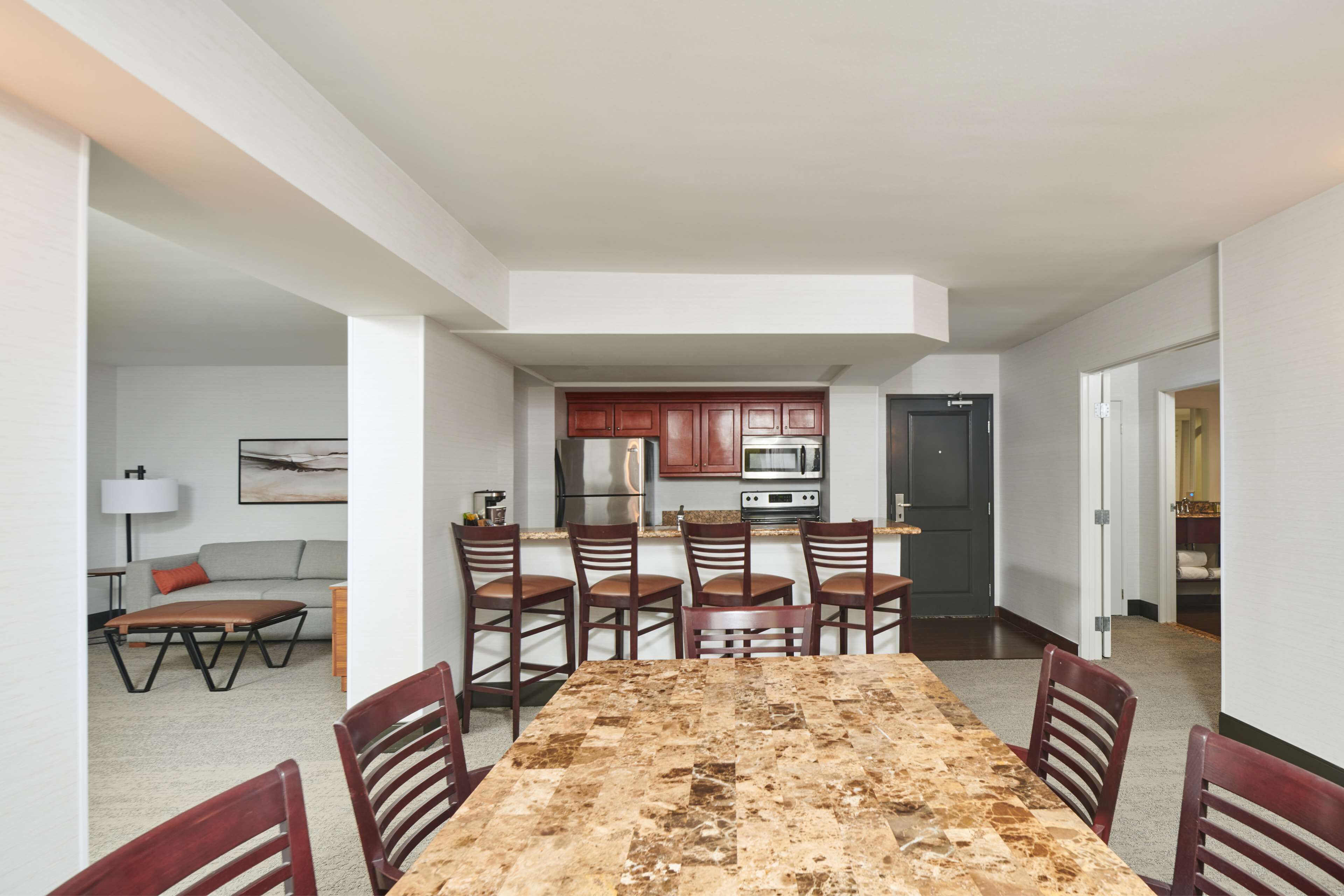 A dining area with a large table and chairs, a kitchen with dark cabinets and appliances, and a living area with a sofa and lamp.