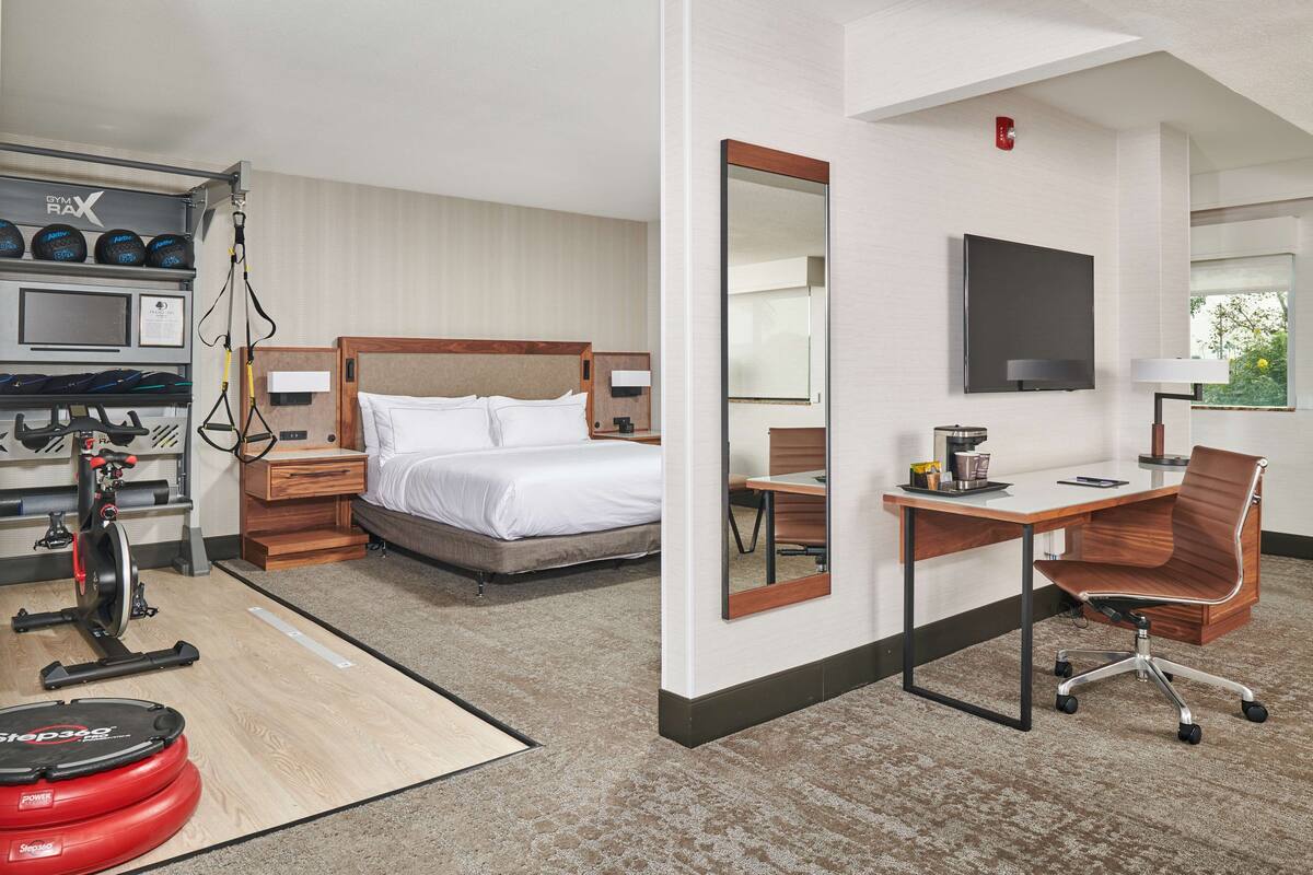 A hotel room with a bed, a desk, a chair, a TV, a mirror, a treadmill, and a set of exercise balls.